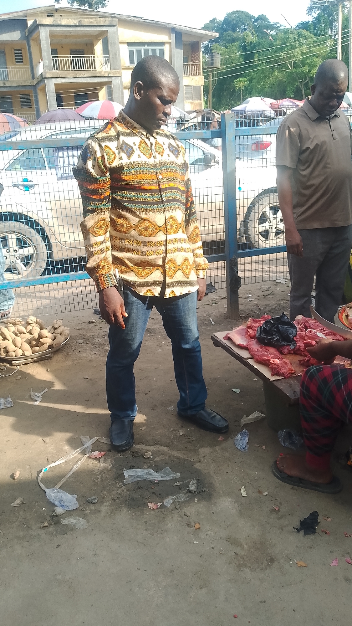 The image captures a street scene in Warri, Nigeria, likely a marketplace or roadside area. A man in a patterned long-sleeved shirt and jeans stands next to a table displaying raw meat, possibly for sale. A woman, dressed in checkered trousers and slippers, is seated near the meat display, perhaps the vendor. Another man stands nearby, wearing a brown shirt and trousers.
There's a basket of what appears to be yams or potatoes to the side. A metal fence and parked cars are visible in the background, suggesting a semi-urban environment. Litter is scattered on the ground, indicating a lack of sanitation. The overall impression is a snapshot of daily commerce in a typical Nigerian urban setting.