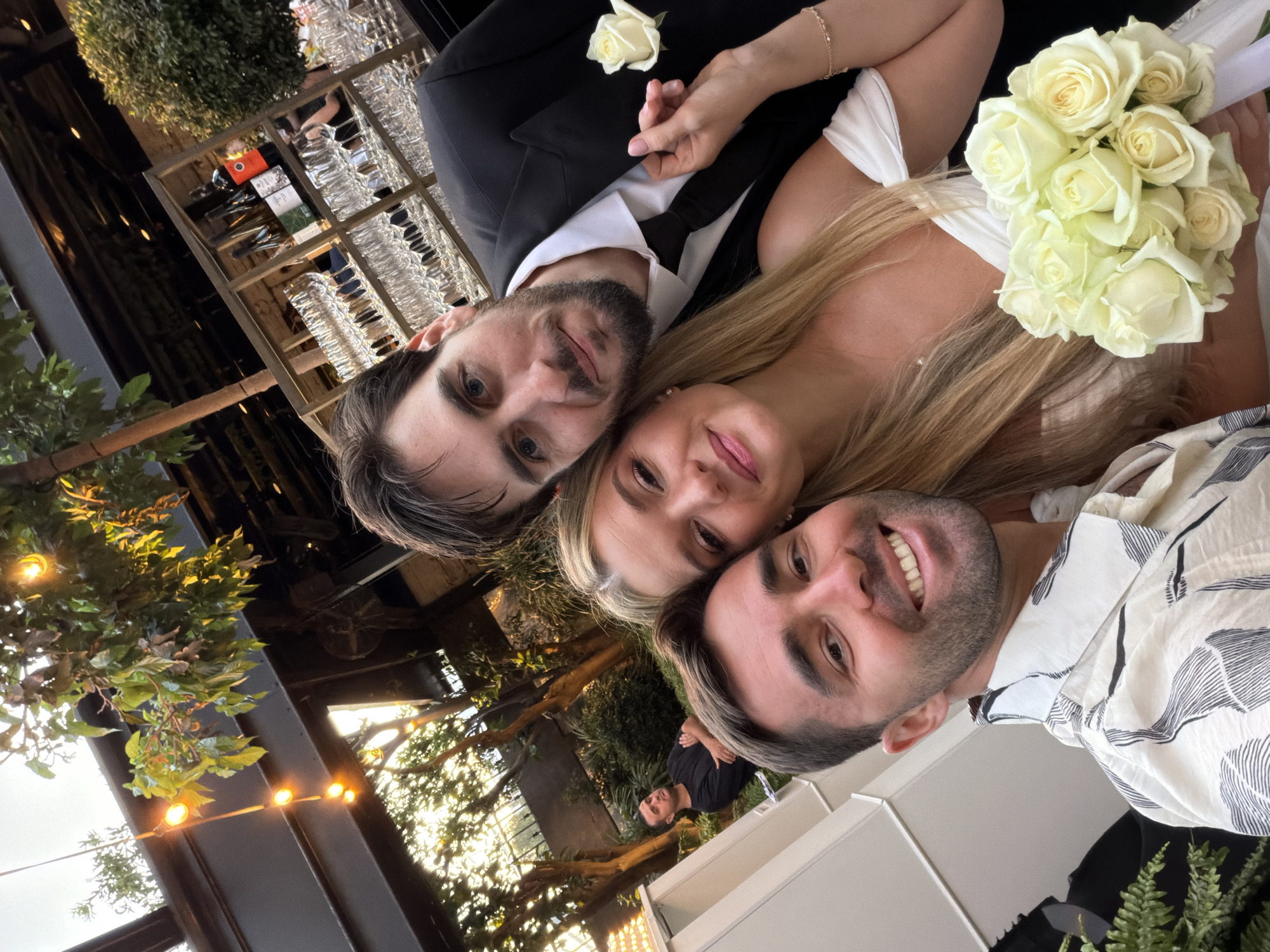 A low-angle shot shows three smiling people, likely at a wedding, in Tel Aviv, Israel. A fair-skinned man in a black suit and tie, with a white rose boutonniere, is to the left of a fair-skinned woman with long blonde hair, wearing a white dress and holding a bouquet of white roses. To their right is another fair-skinned man in a white shirt with a black leaf pattern. In the background are string lights and greenery against a glass and steel building. Wine glasses are visible on a rack behind the man in the suit.