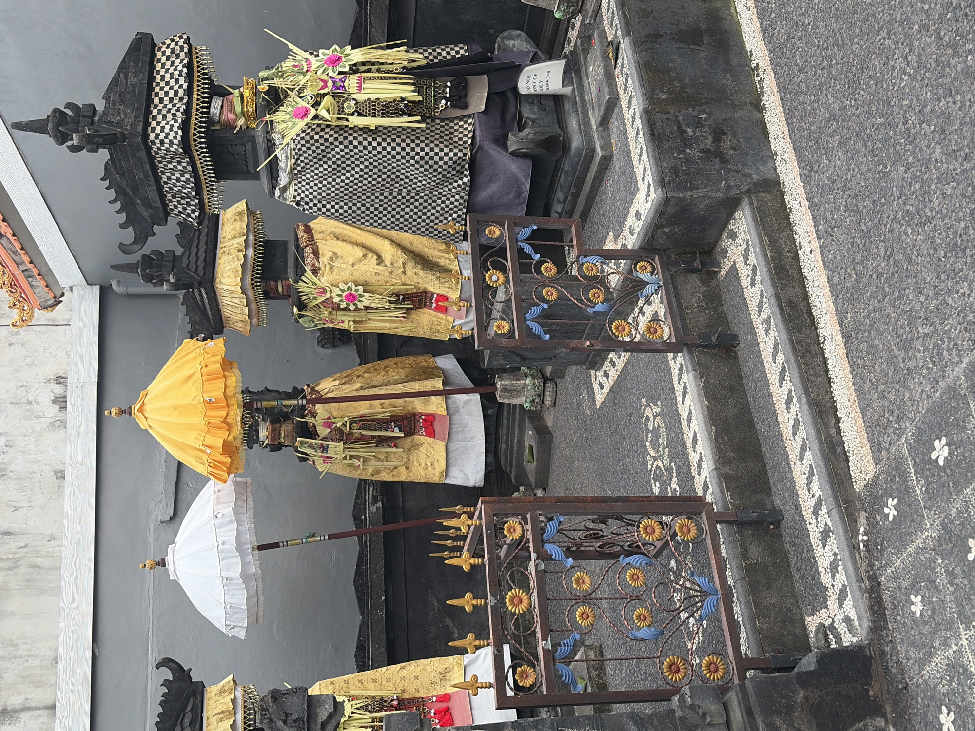The image shows a collection of Balinese offerings in what appears to be a temple or sacred site in North Kuta, Indonesia. There are at least four elaborately decorated figures or shrines, draped in golden cloth and adorned with woven palm leaves and pink floral decorations. Two of the figures are topped with ornate black stone structures resembling miniature temples. In front of these, there are decorative metal fences with floral motifs.
One figure is accompanied by a white umbrella, and another by a bright yellow umbrella, both with decorative tops. The ground is paved with dark pebbles and white stones arranged in patterned borders, and there are some steps leading up to the area where the figures are situated. A sign with text is visible near one of the figures. The background is a plain grey wall. The overall impression is one of a religious or ceremonial setting, likely a public space given the presence of the sign.