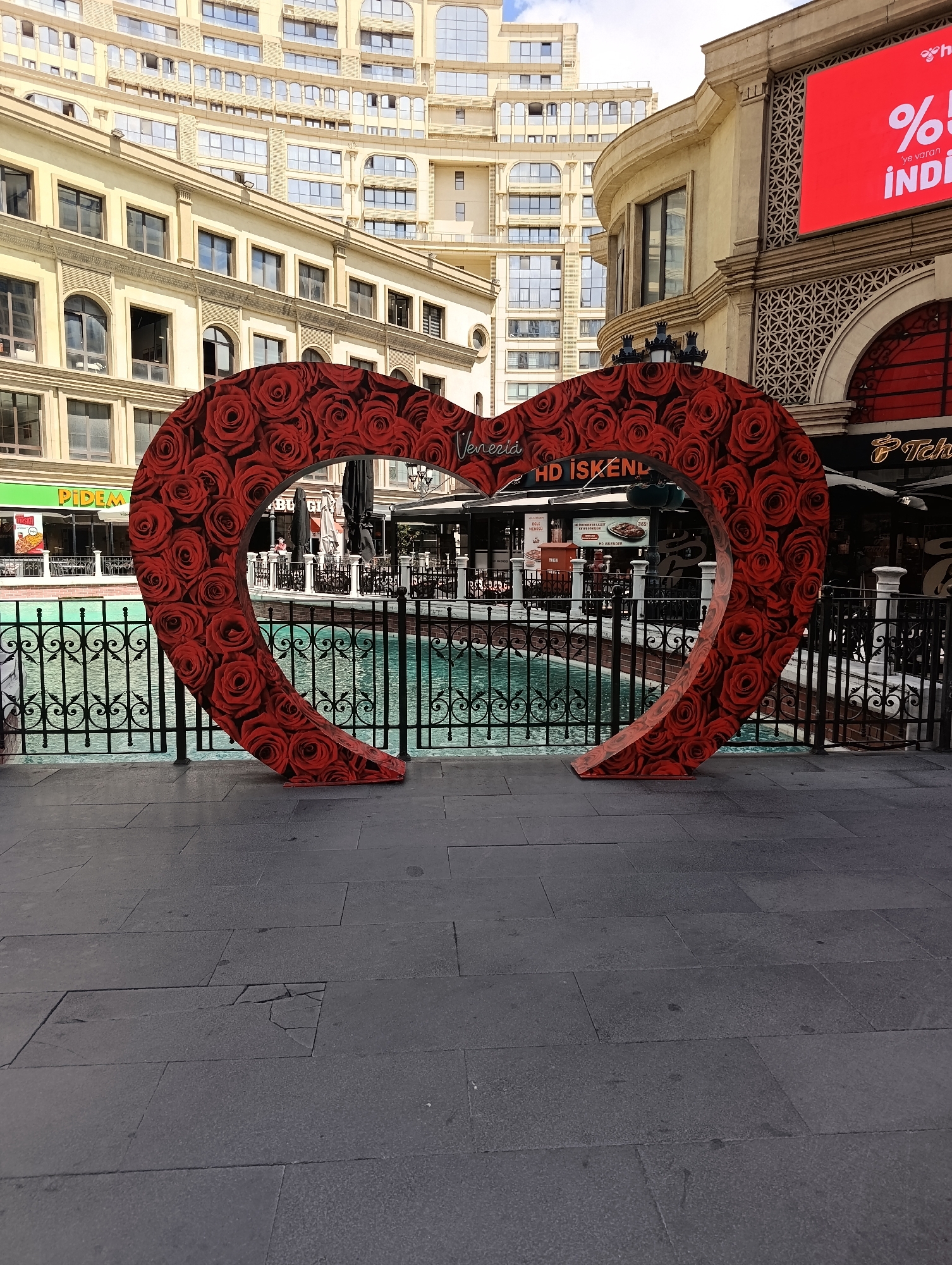 The image depicts a decorative, oversized heart sculpture covered in red roses, situated outdoors in front of a canal. Behind the sculpture, a black wrought-iron fence separates the walkway from the turquoise water. In the background, there are several buildings. The closest building features shops and restaurants, including one with a green sign that reads "PIDEM" and another with a sign for "Tchibo". To the right, a large red digital billboard displays text, including "%" and "İNDİ". Further back, a tall, multi-story building with numerous windows is visible. The ground in the foreground is paved with large, dark gray rectangular stones, one of which is cracked. The scene is illuminated by daylight, suggesting it was taken during the day. This is an outdoor scene at the Venezia Mega Outlet mall in Gaziosmanpaşa, Istanbul, Turkey, a place known for its Venice-themed architecture and canals.