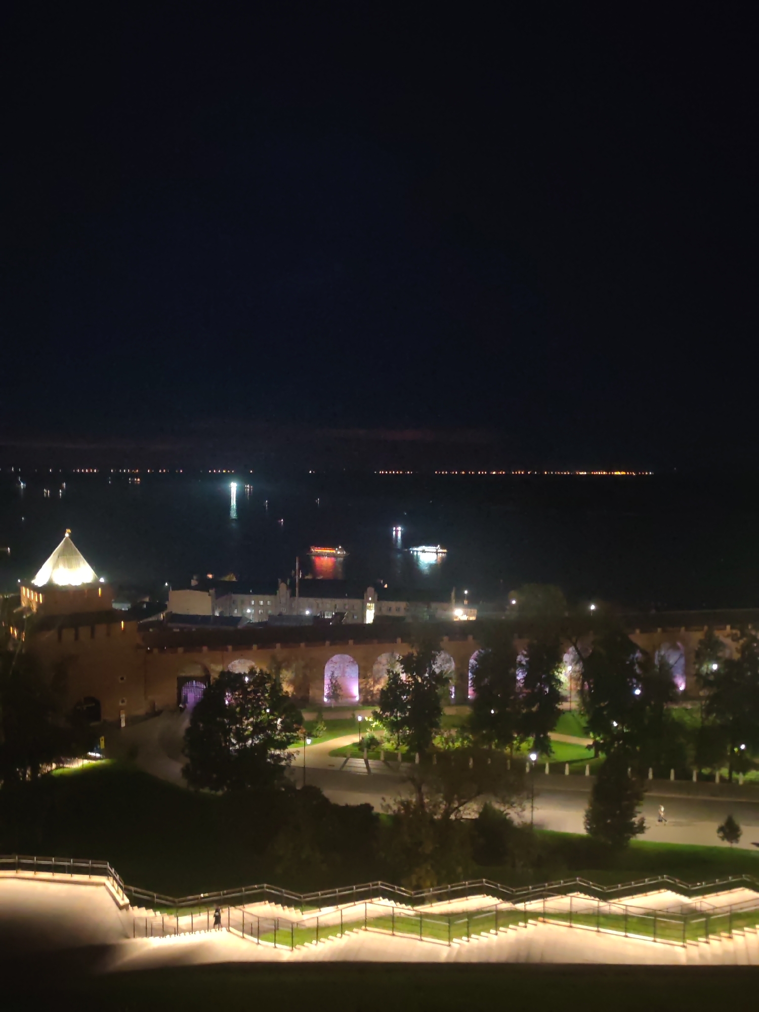 The image depicts a nighttime view of the Nizhny Novgorod Kremlin and the Volga River. The Kremlin walls, illuminated by spotlights, stretch across the middle ground. A conical tower, also lit, is visible on the left side of the Kremlin. In the foreground, a brightly lit staircase with railings winds down a grassy slope. The Volga River is in the background, with lights from boats and distant shorelines reflecting on the water. The sky is dark and clear. Two individuals are visible on the illuminated staircase.