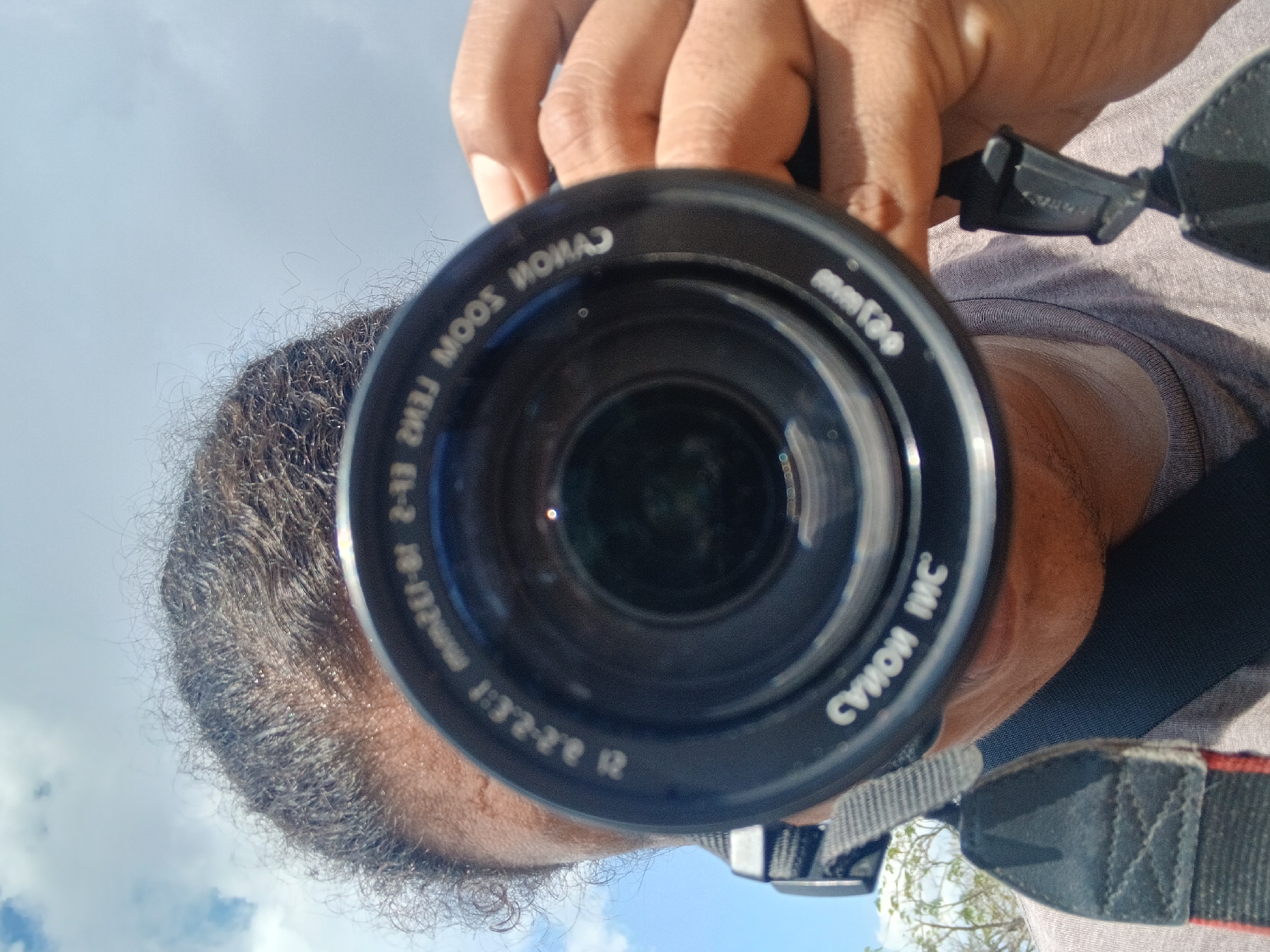 An upward-angled self-portrait or reflection shot captures a person holding a Canon camera, with the lens prominently facing the viewer. The person's hands grasp the camera, and the side of their head, featuring dark, curly hair and an ear, is visible to the left of the lens. They are wearing a light-colored, possibly grey, top.
The camera lens, a "CANON INC. ZOOM LENS EF-S 18-135mm 1:3.5-5.6 IS" with a "Ø67mm" filter thread, has its mirrored text clearly legible across its barrel. The lens reflects some bright light, indicating a sunny environment. A black camera strap with red stitching or accents is visible, draped over the person's shoulder/neck area.
The background shows a bright, clear blue sky dotted with fluffy white clouds, typical of daytime weather. In the lower right corner, a hint of green foliage suggests the presence of trees or bushes, placing the scene outdoors. The high angle and bright lighting create a dynamic and somewhat abstract composition, focusing on the act of capturing an image. The setting is consistent with an outdoor location in Ciudad Guayana, Venezuela, during daylight hours.