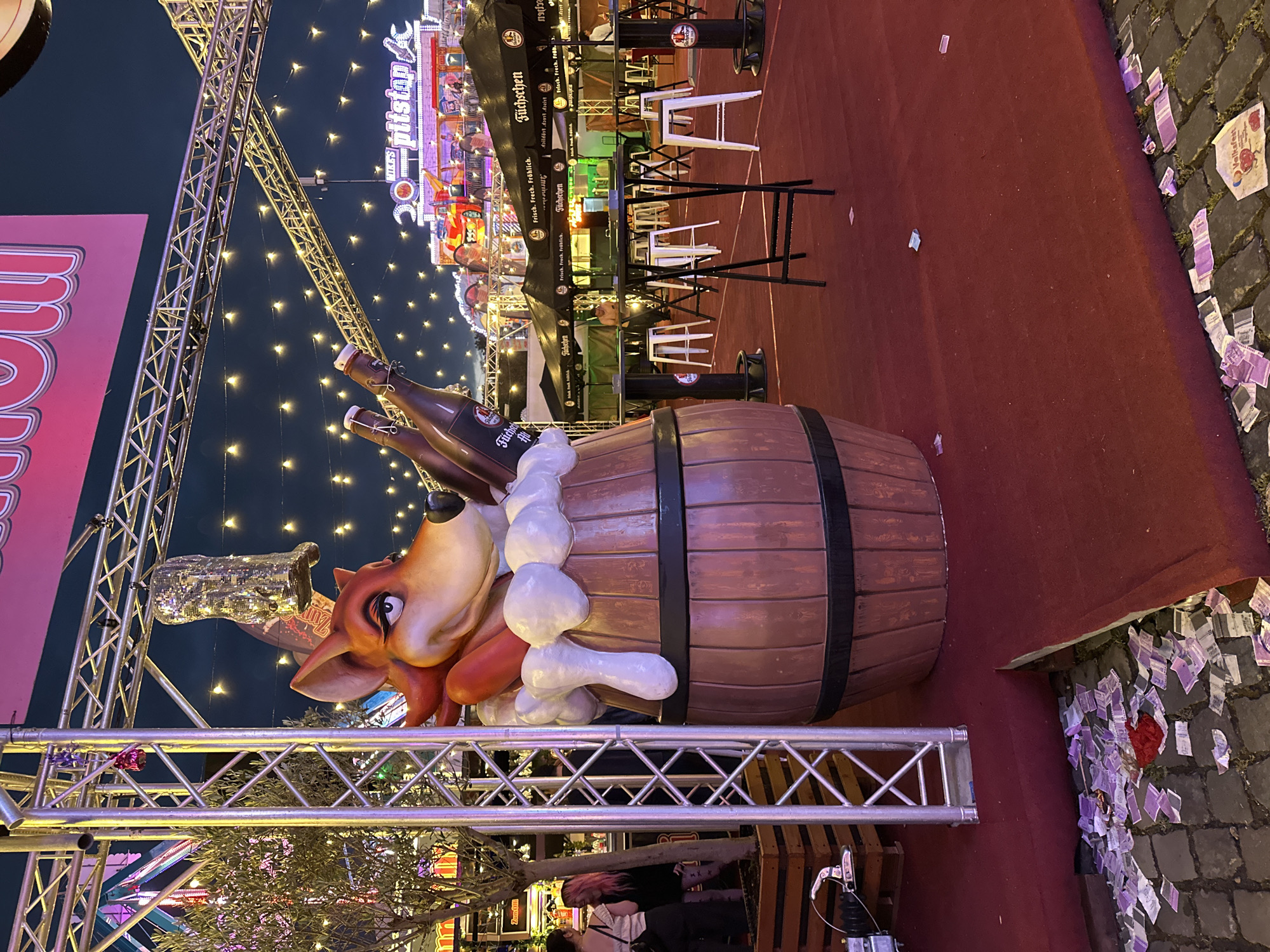 A festive scene unfolds at the Düsseldorf Volksfest. A large, wooden barrel sculpture, overflowing with what appears to be foamy beer, is adorned with a cartoon fox character wearing a festive hat and holding two bottles of beer. The sculpture is situated on a red carpet, a stark contrast to the cobblestone ground scattered with discarded tickets and wrappers. In the background, various food stalls with black awnings are visible, bearing the "Füchschen" logo. A Ferris wheel and other amusement park rides are illuminated in the distance, suggesting a lively atmosphere. The sky is dark, indicating it is evening, and numerous string lights are draped across the area, casting a warm glow. A few people are visible in the background, some interacting near a tree, while others are seated at high-top tables and stools.
