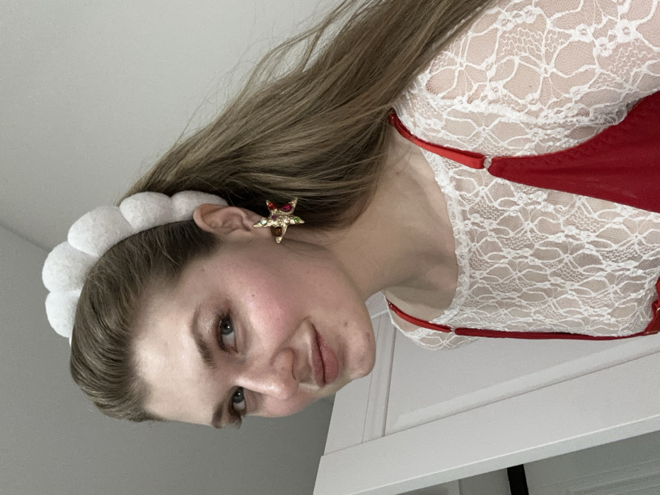 A woman faces the camera, smiling slightly. She has light brown hair and wears a white, puffy headband. A gold, jeweled star earring adorns her ear. She is dressed in a white lace top with red straps and accents. The background consists of a white wall and the corner of a white cabinet or door. The image is slightly rotated.