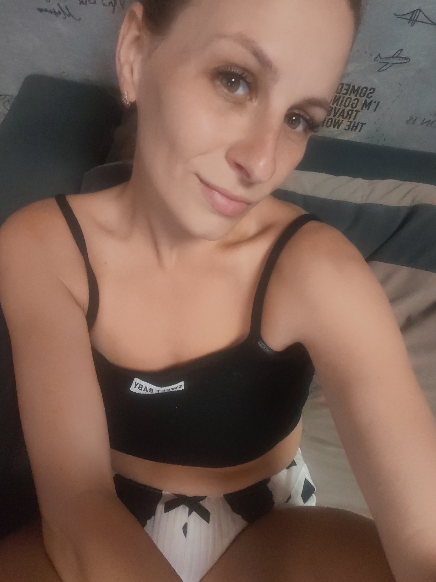 This is a medium shot selfie of a young woman, taken indoors. She is wearing a black bralette with the text "SWEET BABY" visible on the band, and white shorts with a black pattern. She has light skin, brown eyes, and her hair is pulled back. She is looking directly at the camera with a slight smile. The background appears to be a bedroom, with bedding and a wall with some text and drawings. One of the drawings is of an airplane, and the text appears to be in English and possibly Russian. The overall mood is relaxed and casual. There is no indication of the time of day or weather. The location is identified as Mezhdurechensk, Russia.