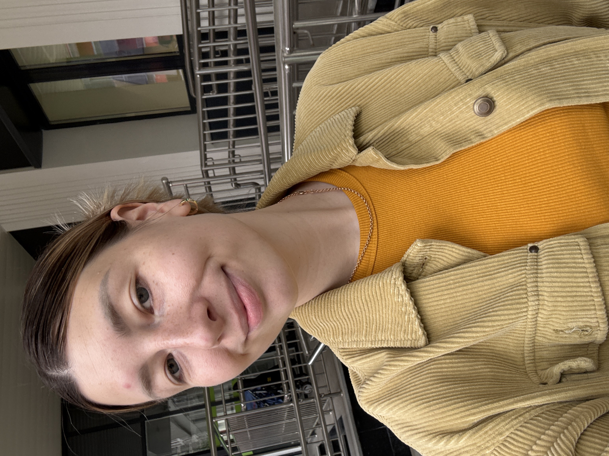 A portrait-style image captures a young woman looking directly at the camera with a gentle, slightly playful smile, her head tilted. She has light skin, dark eyebrows, and light-colored eyes. Her brown hair, with subtle lighter streaks, is pulled back. A small red blemish is visible on her forehead, and a few freckles adorn her face. She wears a delicate gold chain around her neck and a small gold hoop earring on her visible right ear.
The woman is dressed in a ribbed, orange-yellow top, layered with a light yellow or beige corduroy jacket. The jacket features prominent vertical ribs and metallic snap buttons.
The background reveals a modern indoor public space, likely in Moscow, Russia. On the right, blurred metallic grates or railings are visible, suggesting an architectural structure like an escalator or a barrier. Further back, reflections of colorful objects or lights are discernible in what appear to be windows or glass panels. The lighting indicates an indoor setting, possibly during the day or a well-lit evening. There is no visible text in the image.
