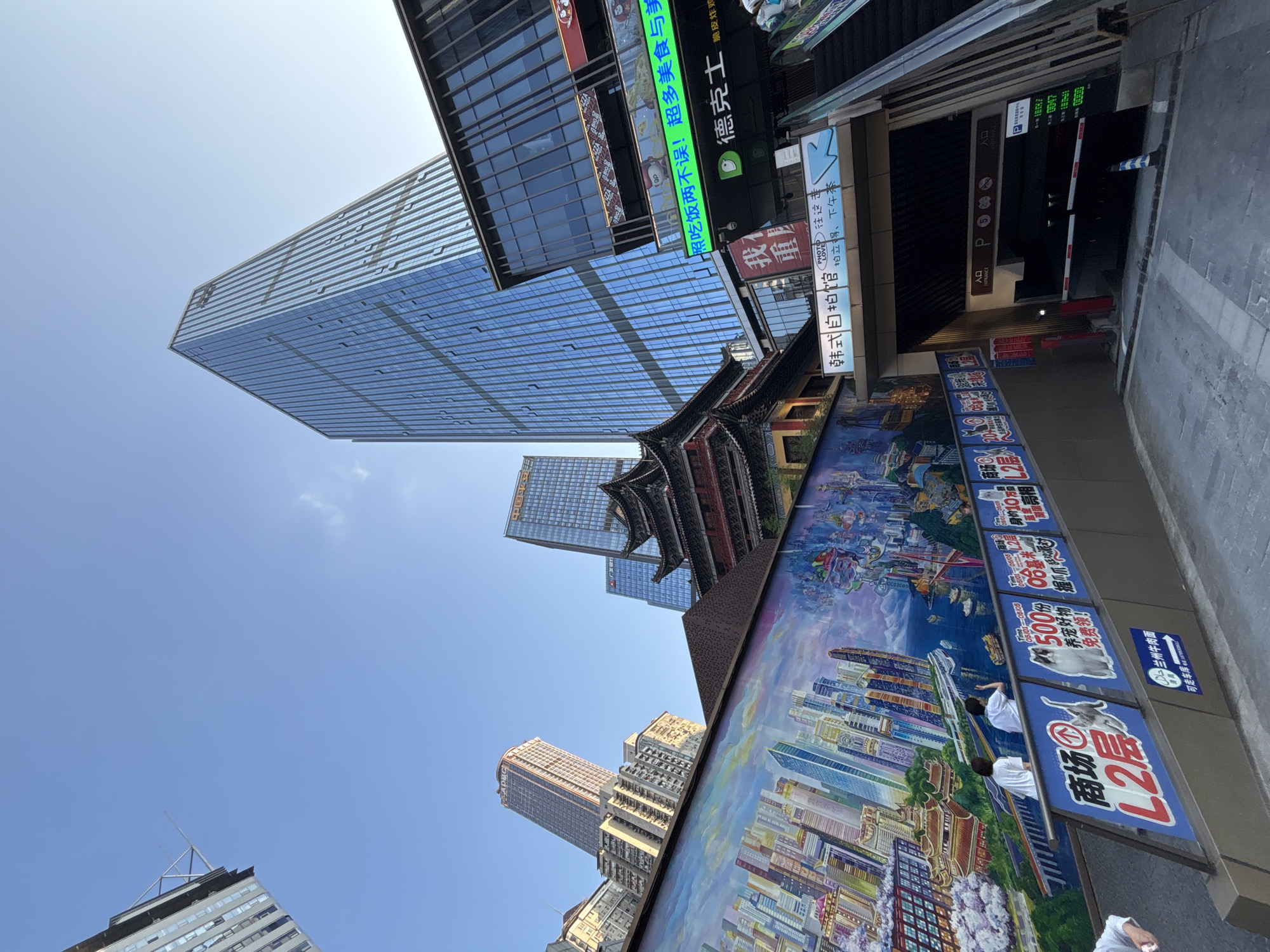 This is a low-angle outdoor shot of a bustling urban scene in Yuzhong District, China. A vibrant mural depicting a futuristic cityscape with skyscrapers and traditional architecture dominates the left side of the frame. Two individuals in white shirts are visible near the mural, one appearing to wave. Several signs with Chinese text are posted along a railing, some featuring images of animals and promotions. In the background, modern glass skyscrapers and a traditional tiered roof building rise against a clear blue sky. An escalator leads into an underground parking garage entrance, marked with "ENTRANCE" and "P" symbols. The overall atmosphere suggests a lively commercial and cultural area.