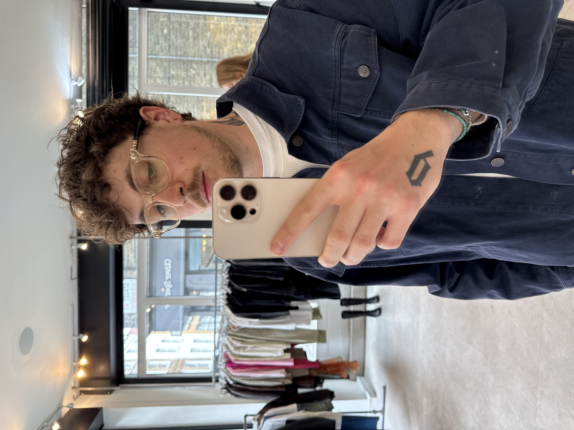A young man takes a mirror selfie inside what appears to be a clothing retail store in London, United Kingdom. The image is rotated 90 degrees counter-clockwise.
The subject is a man with dark, curly hair, a short beard, and light-framed eyeglasses. He is wearing a dark blue, possibly corduroy or ribbed, button-up jacket over a white undershirt. His right hand, holding a light-colored smartphone (likely an iPhone with a triple camera setup), features a distinct, bold, Gothic-style tattoo of the number "6" or letter "G". A turquoise beaded bracelet and a silver chain are visible on his wrist, and further outline tattoos can be seen on his neck. He has a nose piercing and an ear piercing.
The setting is a brightly lit indoor space with white walls and a light-colored floor. Reflected in the mirror behind him are multiple racks of clothing, displaying various garments in different colors, indicating a boutique or clothing store environment. A large window provides a view of an urban landscape outside, showing brick buildings under a daytime sky. Wall-mounted spotlights illuminate the interior.
Notable details include text on the jacket buttons, partially visible as "DENIM CO". Reflected in the window are reversed texts that appear to be "OTRO-2900" and "2MEER", likely signs from outside the establishment. The man's expression is neutral, captured in the act of taking a mirror photo.