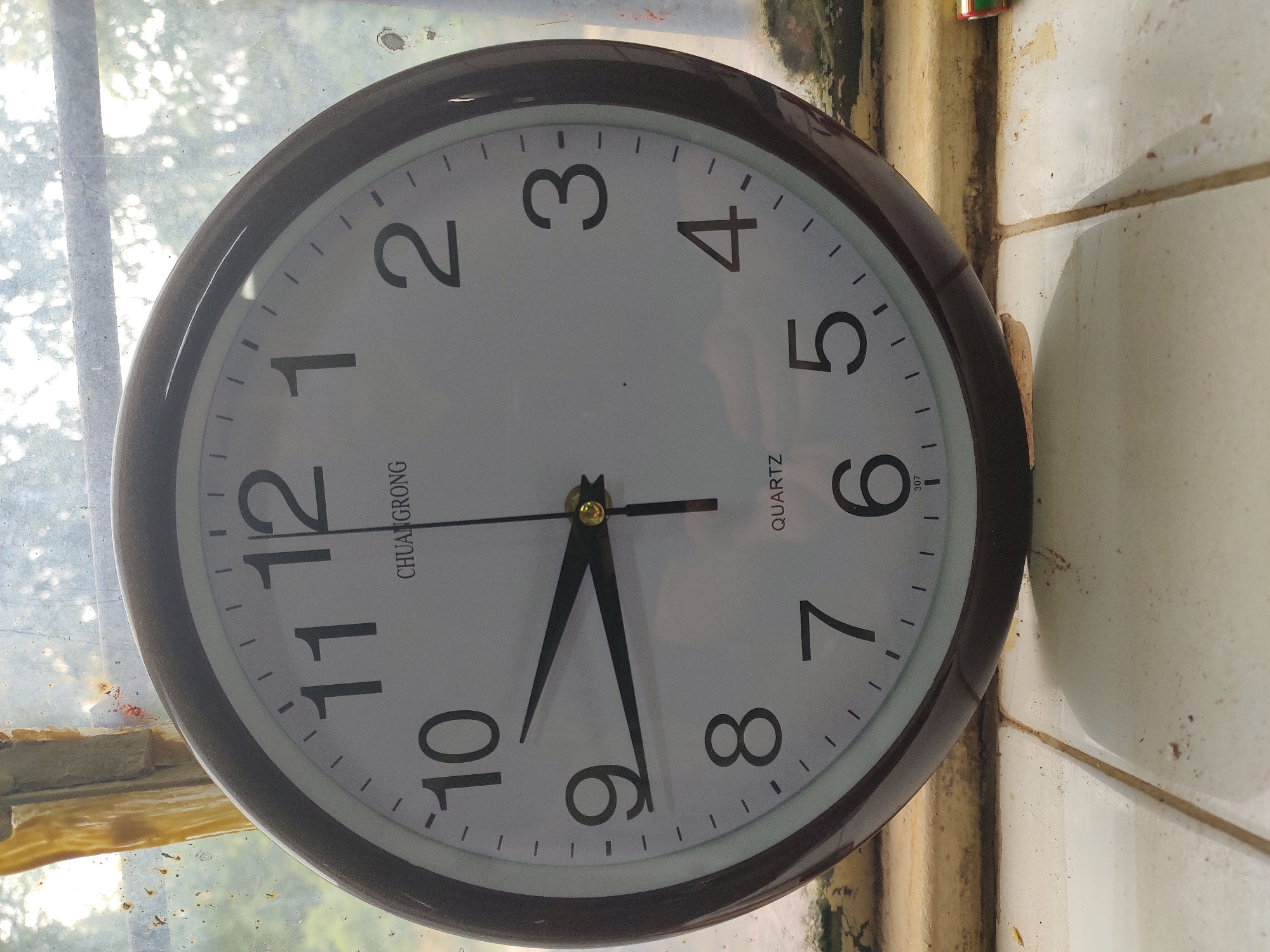 A dark-rimmed, circular analog clock with a white face is displayed. The clock has black hour and minute hands, with the hour hand pointing slightly past the 9 and the minute hand pointing to the 10. The numbers 1 through 12 are clearly visible in black. The clock is positioned on a ledge, possibly a windowsill, against a backdrop of a hazy, out-of-focus window with sunlight filtering through. The environment appears somewhat weathered, with visible grime and dust on the ledge and the window frame. The background suggests an outdoor setting with foliage. The clock face reads "CHUANGRONG" and "QUARTZ".