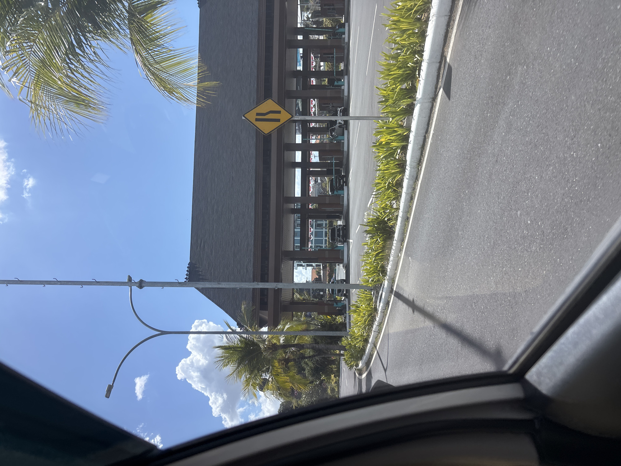 The image, taken from a vehicle on a sunny day in Kuta, Indonesia, shows a road leading towards a building with a dark, sloped roof supported by wooden pillars. The building appears to be a covered structure, possibly an entrance or a waiting area. In front of the building, there are multiple lanes marked with barriers. A yellow diamond-shaped road sign with black arrows indicating the road narrowing is visible on the right side of the road. Palm trees and lush green plants line the roadside. The sky is blue with scattered white clouds. Several cars are visible within the covered structure. The perspective suggests the photo was taken while driving past this location.