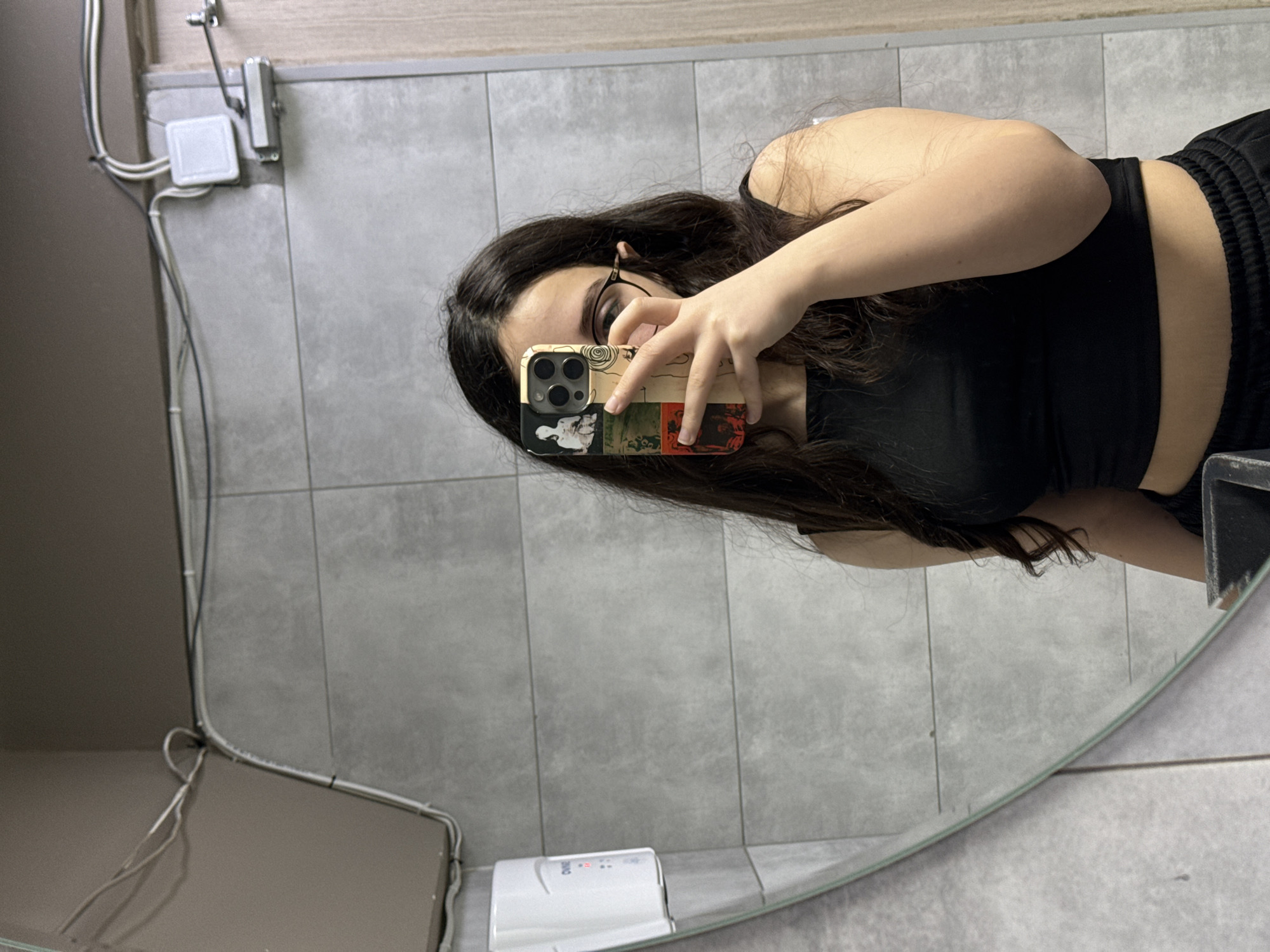 An individual is captured in a mirror selfie, obscuring their face with a smartphone held in their right hand. The person has long, dark, wavy hair and wears dark-rimmed glasses, a black sleeveless crop top, and black athletic-style pants. The smartphone features a multi-panel case with various black, white, green, and red designs.
The environment is an indoor space, consistent with a public or commercial restroom. The walls are predominantly covered with light grey square tiles. Above the tiled section, the upper wall and ceiling are a plain light brown or beige color. In the background reflection, a white wall-mounted device, possibly a hand dryer or soap dispenser, bearing the visible text "OWN" and indicator lights, is present on the left. Exposed white and grey electrical conduits with cable ties run along the upper wall, connecting to a white square junction box. On the right, a wooden door frame or partition is visible with a silver-colored door closer mechanism attached near its top. The bottom edge of the mirror shows a dark, possibly metallic or tiled, frame or countertop.
The specific building or street address within Yerevan, Armenia, is not identifiable from the visible details. The setting is an indoor public or commercial restroom facility, characterized by its tiled walls and specific fixtures, consistent with establishments found in an urban environment like Yerevan.