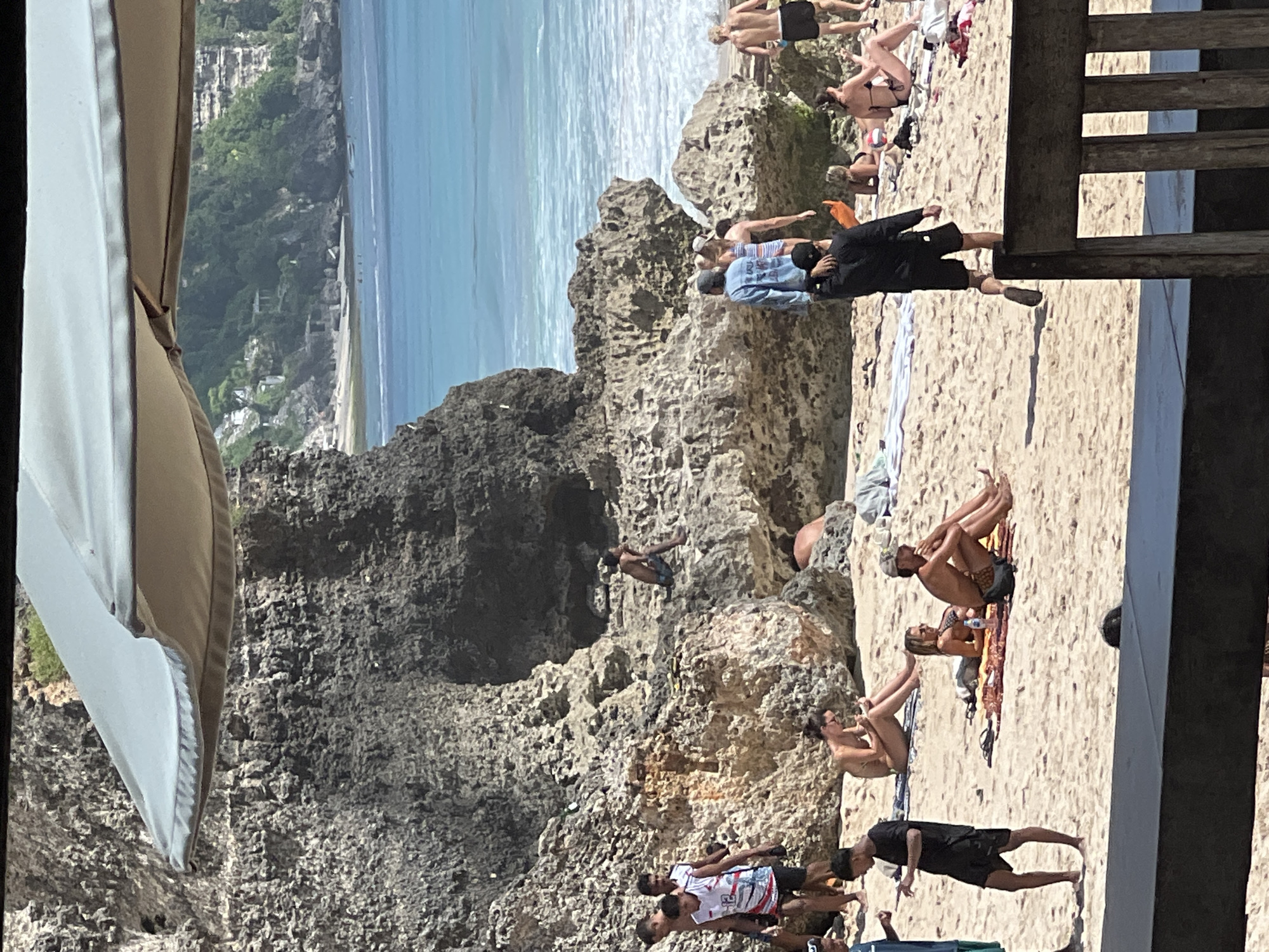 The image depicts a crowded beach scene in Kuta Selatan, Indonesia, featuring a rocky cliff formation and a view of the ocean. People are scattered across the sandy beach, engaging in activities such as sunbathing, walking, and interacting with each other. Some individuals are positioned on the rocks. The scene is partially framed by structures, including a light-colored fabric canopy or awning on the left and wooden beams on the right. The ocean is visible in the background, exhibiting a gradient of light blue colors. A densely vegetated hillside can be seen on the left side of the frame.
