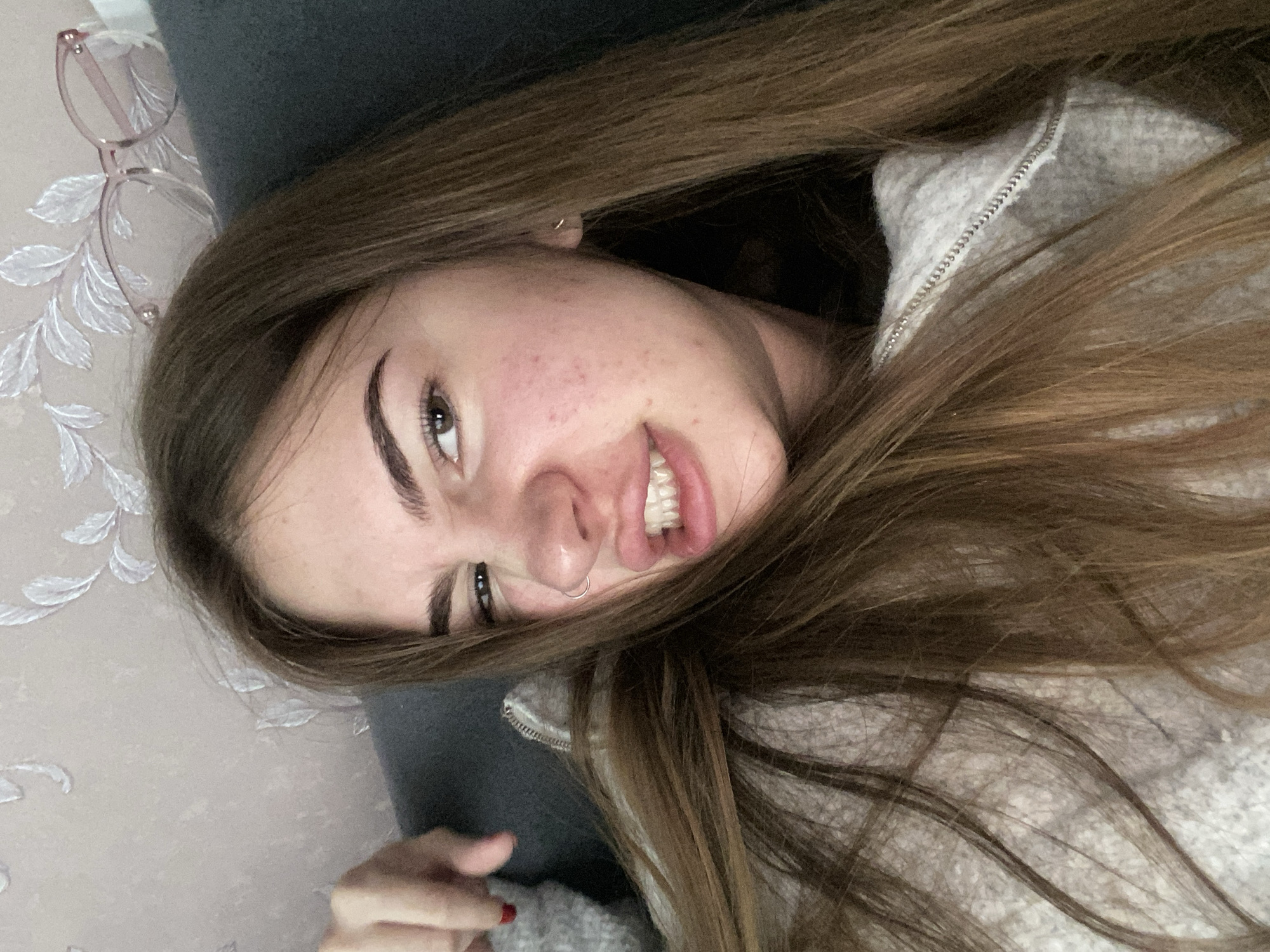 A young woman is prominently featured in a close-up, rotated image, appearing to be lying down indoors, possibly on a bed or sofa, while looking towards the camera. She has long, straight to slightly wavy brown hair that frames her face and spreads around her. She wears a playful, slightly mischievous smile, revealing her teeth, and her dark eyes are directed at the viewer. Her skin is fair, with some redness visible on her cheeks. A small silver hoop nose ring adorns her right nostril. She has well-defined dark eyebrows and a small, subtle hoop earring is visible on her right ear.
She is dressed in a light grey, fuzzy or fleece-like hooded garment, with a silver zipper partially visible around her neck and chest area. Her left hand, with a hint of red nail polish on her fingers, is partially visible towards the bottom of the frame.
The background consists of a dark grey cushion or pillow positioned behind her head. Behind that, a light-colored wall with a subtle, raised white floral or leafy pattern adds texture to the setting. A pair of translucent pink-framed glasses with clear lenses are resting on this patterned wall, near her head. The overall impression is one of relaxation and lightheartedness. There is no visible text in the image.