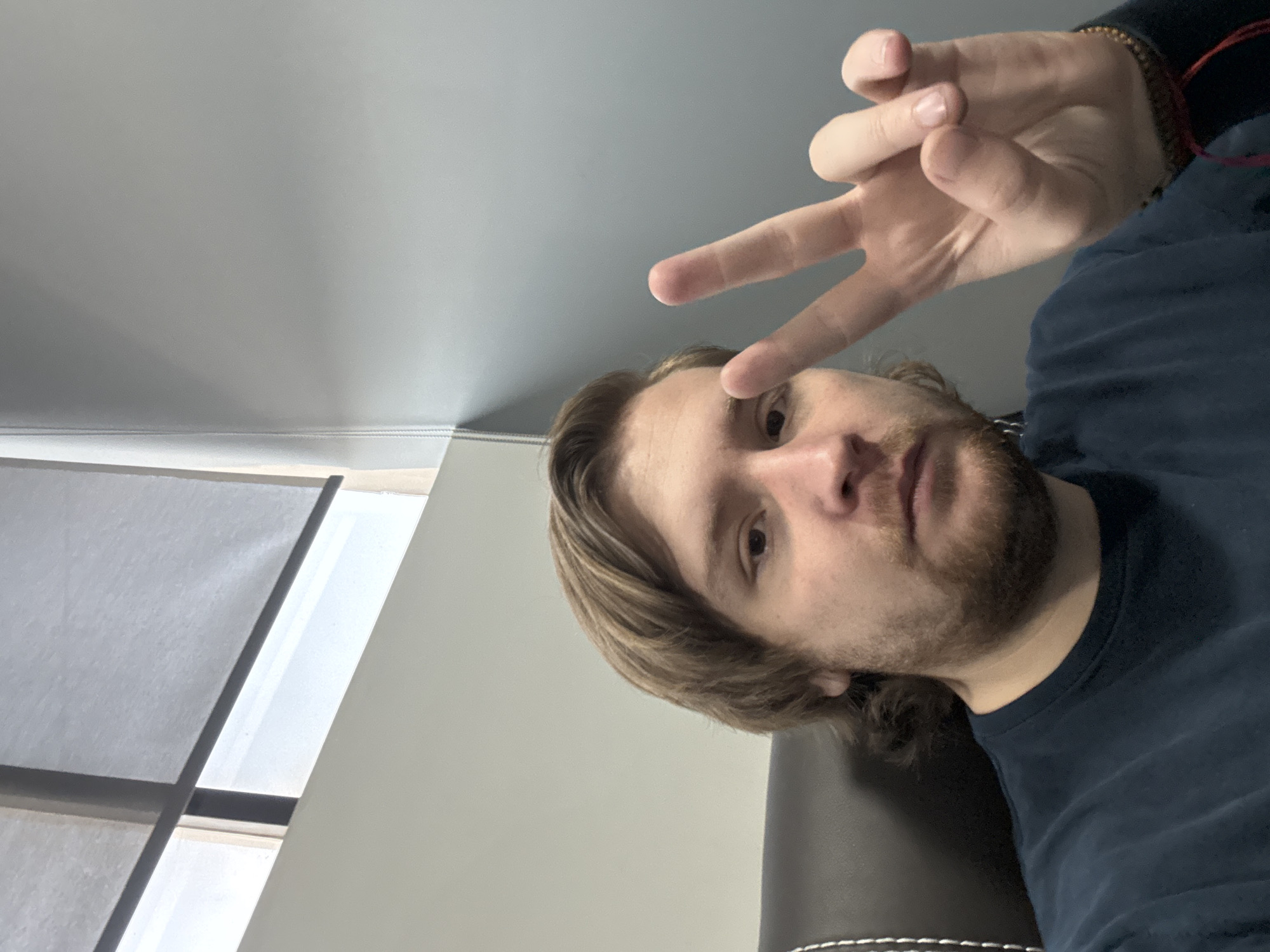 A selfie of a man with light brown hair and a beard, wearing a dark blue shirt and a beaded bracelet, is making a peace sign with his hand. He is sitting in a dark-colored chair with white stitching. To the left, there is a window with white blinds, and above him, there is a plain light-colored wall and ceiling. The location is not discernible from the image. There is no information about the city of Cuautitlán Izcalli, Mexico.