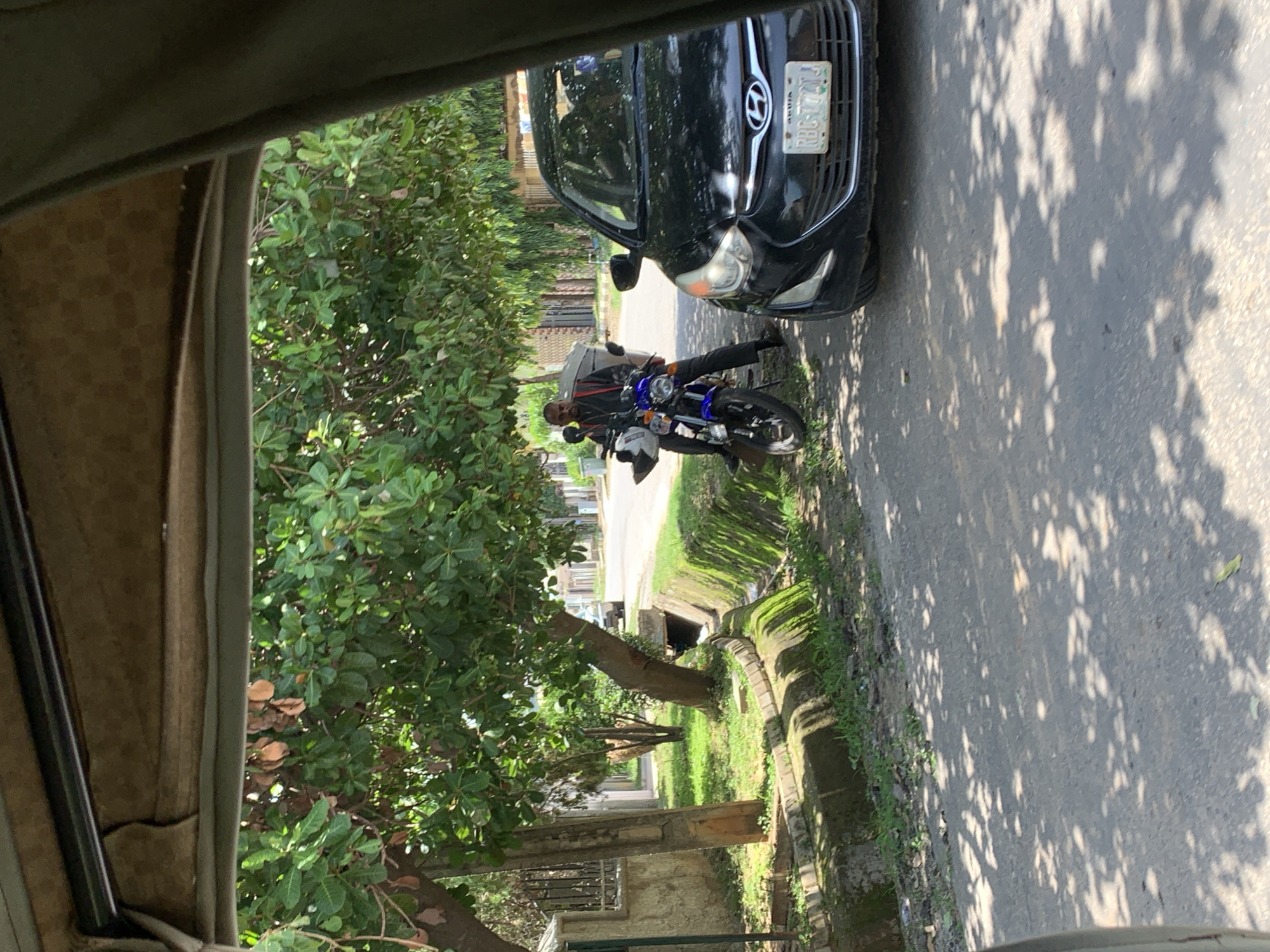 The image depicts a man on a motorcycle parked next to a black Hyundai car. The scene appears to be a street in Abuja, Nigeria, with lush green trees and a drainage ditch visible. The sunlight filtering through the trees casts dappled shadows on the asphalt road, suggesting it is daytime. The man is wearing a dark jacket and trousers, and he is looking forward. The motorcycle is blue and white, and the car has a license plate with the text "KRG 177 JJ". The overall impression is one of an ordinary day, with no clear indication of any particular event or emotion. The perspective suggests the photo was taken from inside a vehicle, possibly a bus or van.