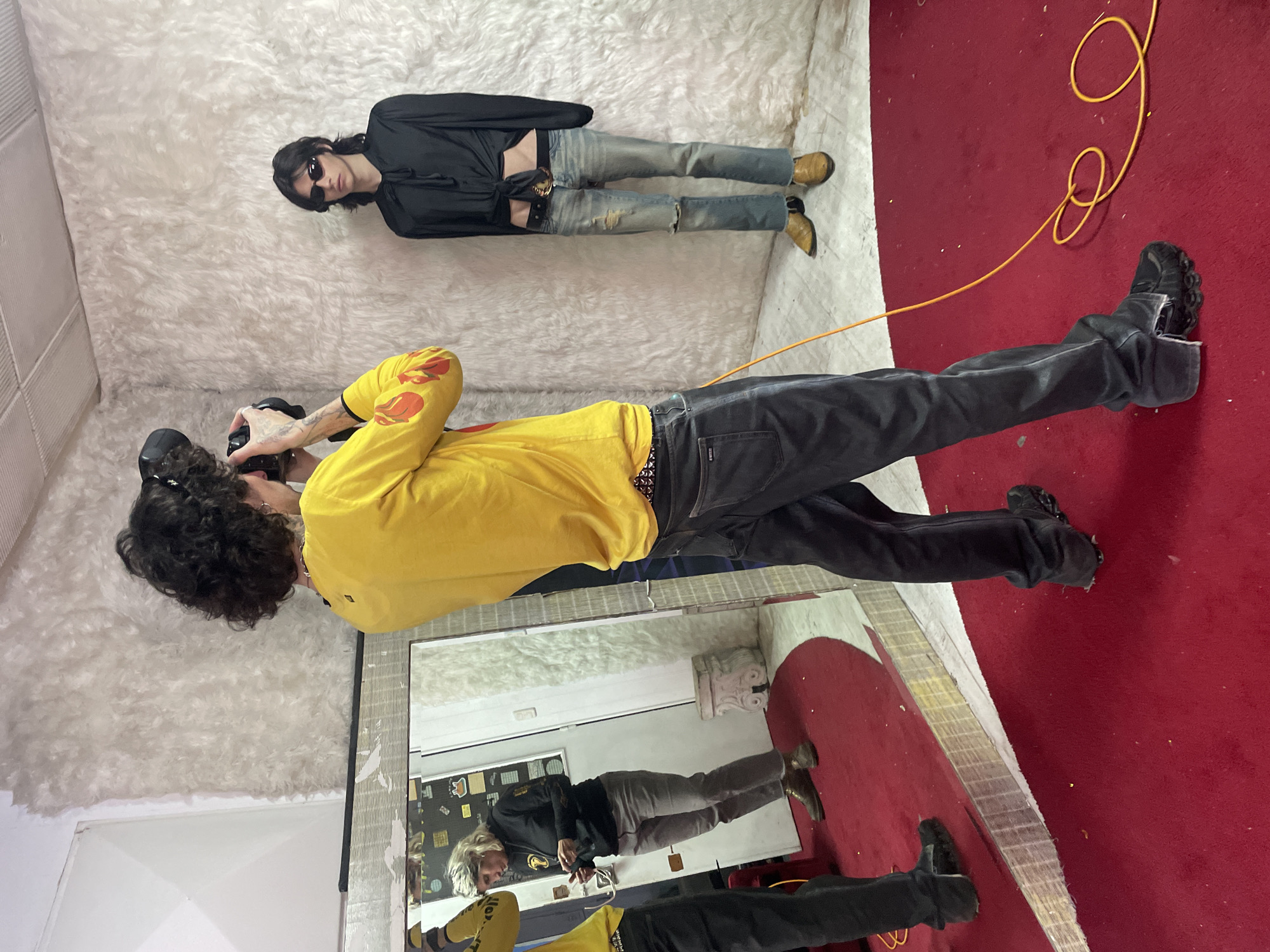 A man in a yellow shirt with flame designs on the sleeves and black pants is taking a picture with a camera, standing in a room with white, textured walls and a red rug. His reflection is visible in a mirror on the wall. Lying on the white, textured surface is a person in a black shirt, blue jeans, sunglasses, and tan boots. An orange electrical cord is on the red rug. The city of Buenos Aires, Argentina is not discernible in the image.