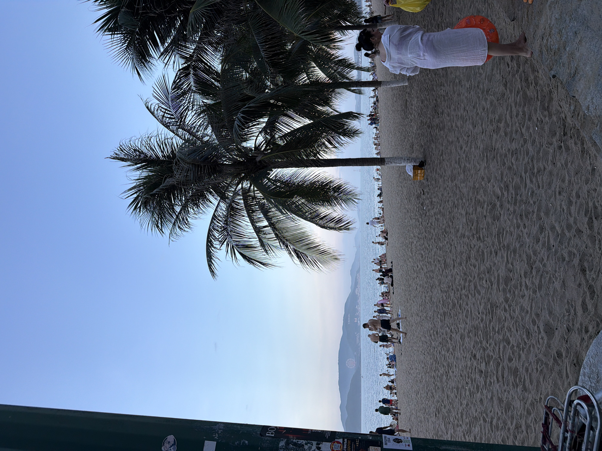 The image depicts a beach scene in Nha Trang, Vietnam, with a clear blue sky overhead and the ocean stretching into the distance. The beach is populated with numerous people, some walking, others sitting or standing, and many are visible near the water's edge. Palm trees are scattered across the sandy expanse, with their fronds providing shade.
In the foreground, a person wearing a white cover-up stands on the sand, holding an orange inflatable ring. To the left, a dark, cylindrical object, possibly a pole or pillar, is visible, adorned with posters. The background features a hazy view of mountains on the horizon, with a Ferris wheel and buildings suggesting a city or resort area. The overall atmosphere is relaxed, indicative of a day at the beach.