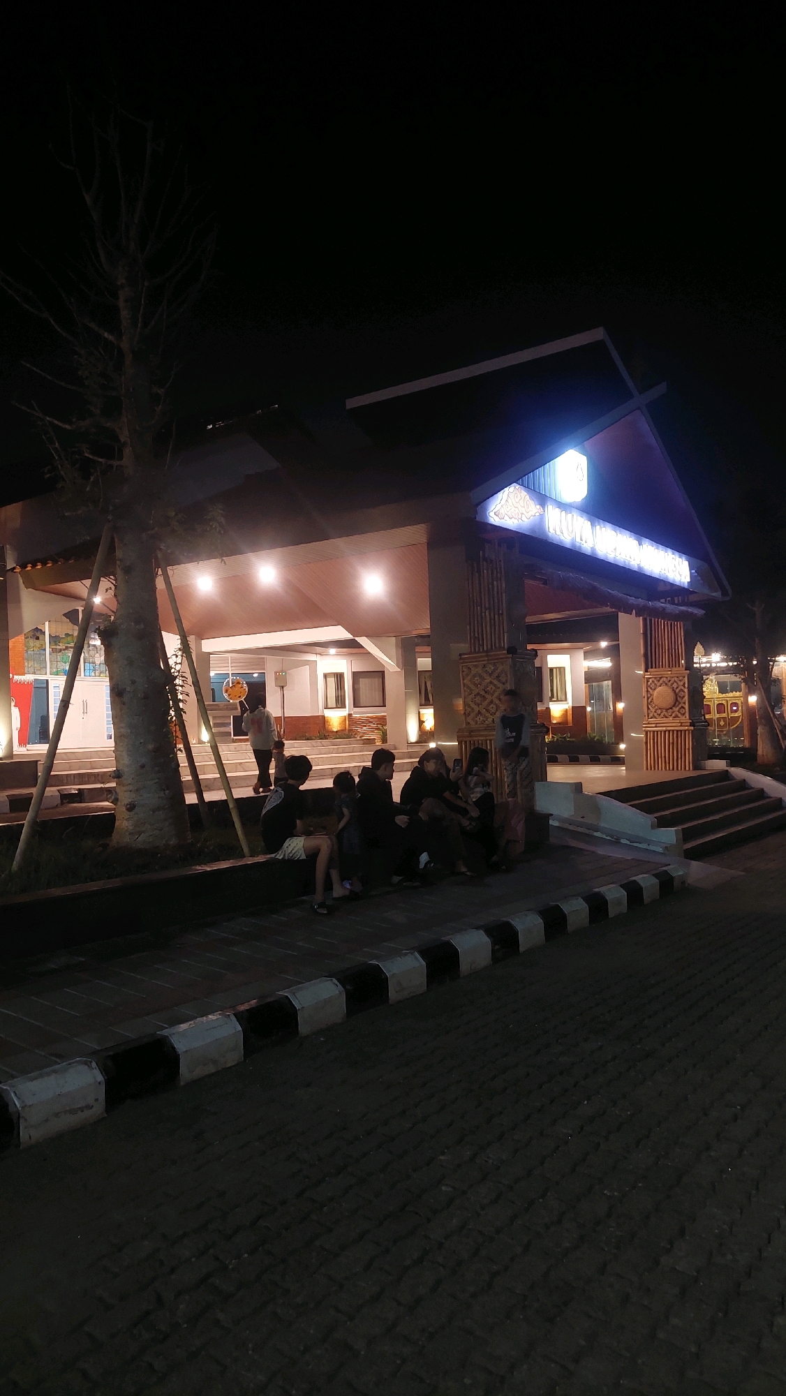 The image depicts the entrance of a building at night, likely a public facility or attraction, situated in Cibinong, Indonesia. The building features a prominent roof structure with illuminated signage in the background, which appears to read "WISATA KULINER KEMANGI" and possibly other text. Several people are gathered, primarily sitting on a low wall or ledge in the foreground and on steps leading up to the building. A tree with bare branches stands to the left of the entrance. The ground is paved with interlocking blocks, and a curb with black and white markings separates it from the pedestrian area. The lighting suggests an evening or nighttime scene, with artificial lights illuminating the building and its surroundings. The overall atmosphere is casual, with groups of people appearing to be socializing or waiting.