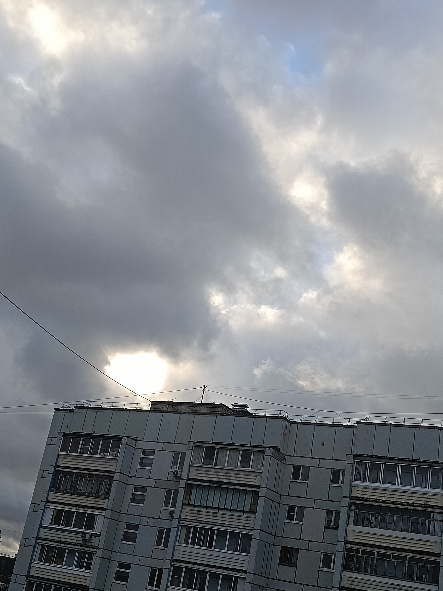The image shows a multi-story apartment building in the city of Chaikovsky, Russia. The building is light blue-gray with rows of windows and balconies. Overcast sky with dark gray clouds. A power line runs diagonally across the upper left portion of the frame. The sun is obscured by the clouds, casting a bright light behind them.