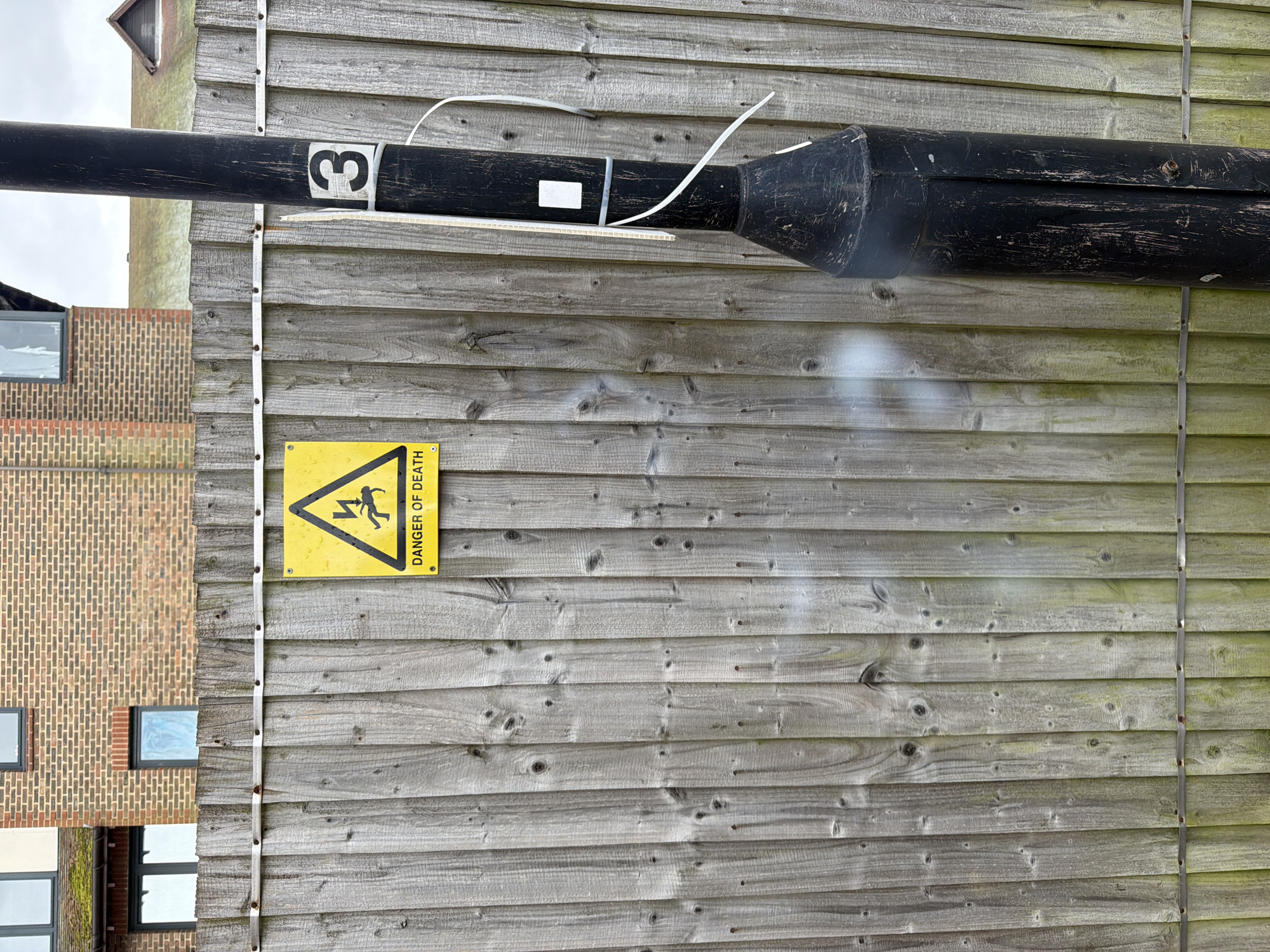 The image shows a wooden fence in Mole Valley, United Kingdom. Affixed to the fence is a yellow triangular "Danger of Death" sign depicting a person being electrocuted. A black, cylindrical object with a number "3" on it, possibly a repurposed piece of industrial equipment, is mounted horizontally along the upper portion of the fence. A building constructed of red and orange brick is partially visible to the left of the fence. The weather appears overcast.