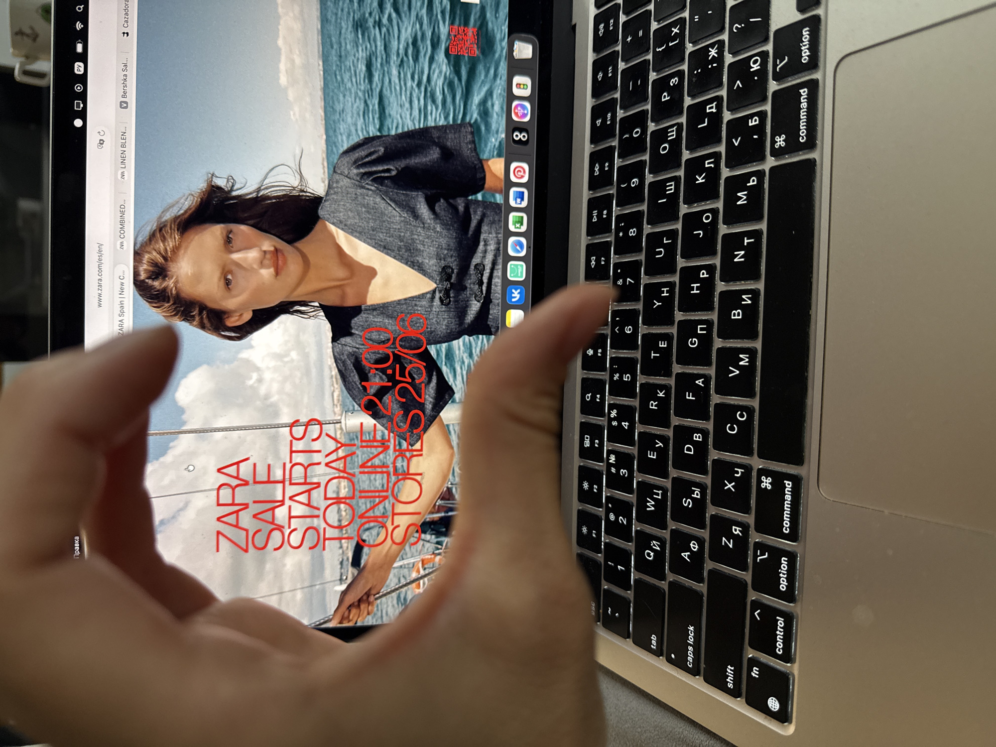 The image displays a laptop screen showing the Zara website with a sale announcement. A hand is partially obscuring the screen. The laptop is on a surface, possibly a table, as indicated by the keyboard. The background of the laptop screen features a model against a sea backdrop. The keyboard shown is a QWERTY layout with some Cyrillic characters superimposed, indicating it is likely set to a Russian language input. The scene does not contain any identifiable elements that would allow for a specific location in the city of Chaikovsky, Russia.