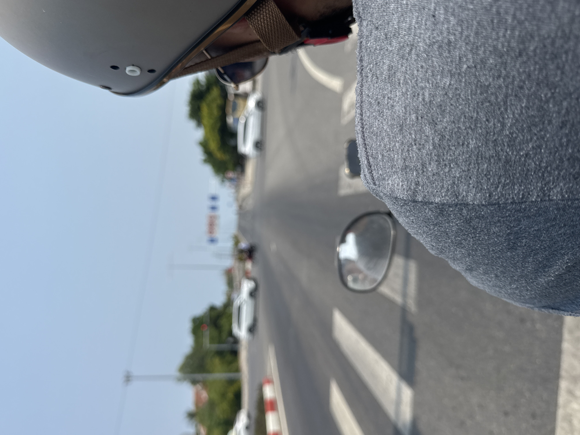 This first-person perspective image captures a scene from a motorcycle or scooter on a clear, sunny day in Da Nang, Vietnam. The viewpoint is looking forward down a road, with the immediate foreground occupied by parts of the rider.
On the left side of the frame, the top and side of a dark grey or black helmet are visible, complete with a brown retention strap. A small white plastic ring and a few small holes are noticeable on the helmet's surface. On the right, a textured grey fabric, likely a sleeve or shoulder of the rider's clothing (such as a jacket or shirt), fills a significant portion of the edge.
In the mid-ground, an oval-shaped rearview mirror is positioned on the right, reflecting blurred sections of the road or sky. The road ahead is paved and features prominent white markings, including a pedestrian crossing with broad white stripes in the foreground and lane lines stretching into the distance.
Along the road, several vehicles, mainly white cars, are present, appearing blurred in the mid-to-far distance. A red and white striped barrier or bollard is visible on the left side of the road near the crossing. The sides of the road are lined with green trees and some indistinct buildings, suggesting an urban or suburban setting. The sky above is bright and clear, indicating fair weather. The background elements are out of focus, creating a sense of depth and directing attention to the immediate surroundings and the path ahead. No discernible text is visible on any signs or objects in the image.