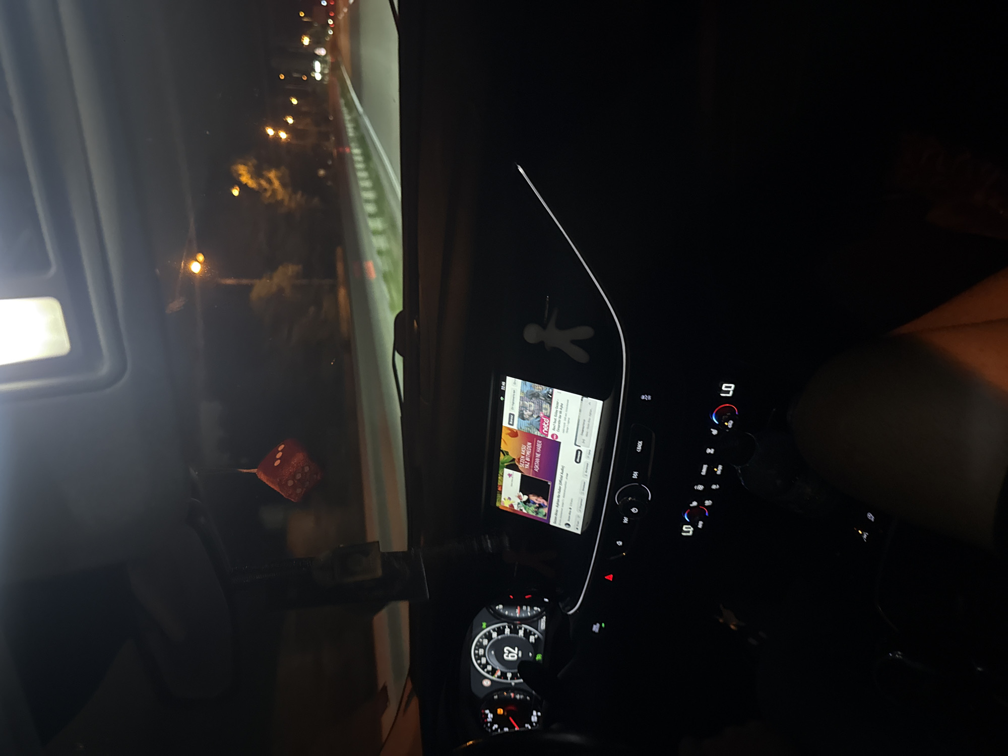 A nighttime scene depicts the interior of a moving vehicle, likely in Varna, Bulgaria, viewing forward onto a multi-lane road. The illuminated dashboard displays a speedometer indicating a speed of 62 (presumably km/h). To the right, an infotainment screen shows a media player interface at 22:46, featuring the artist Sezen Aksu and the song "Aşktan Ne Haber" (Turkish for "What's Up with Love") from "netd müzik". Below the screen, climate controls are set to "LO" on both driver and passenger sides. A red fuzzy dice decoration hangs from the rearview mirror area. A small, white, humanoid figure adheres to the dashboard to the right of the infotainment screen. A hand is partially visible on the steering wheel, indicating a driver. Outside the vehicle, the road is illuminated by a series of warm yellow streetlights, with trees and a road barrier visible alongside. Distant lights suggest other vehicles or urban infrastructure ahead.