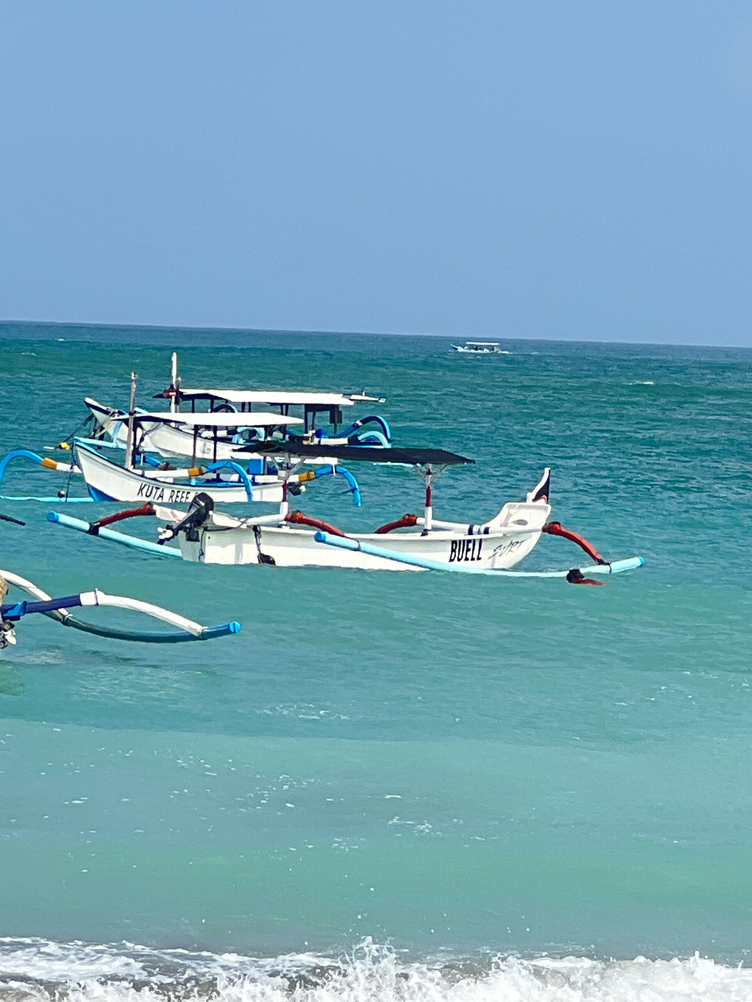 In the city of Kuta, Indonesia, a vibrant scene unfolds on the turquoise waters of the ocean. Several traditional Indonesian outrigger boats, known as jukungs, are anchored or gently bobbing on the gentle waves. One prominent boat, white with blue trim and the name "KUTA REEF" emblazoned on its side, features an outboard motor. Another, also white with blue accents, bears the name "BUELL" and a stylized signature. These vessels are equipped with outriggers for stability, painted in bright red and blue, and some have canopies for shade. In the distance, a smaller boat is visible on the horizon. The clear blue sky and the shimmering ocean create a serene and picturesque setting, characteristic of Kuta's popular beaches, likely captured during the day under sunny conditions. The foreground shows the gentle surf of the ocean meeting the shore.