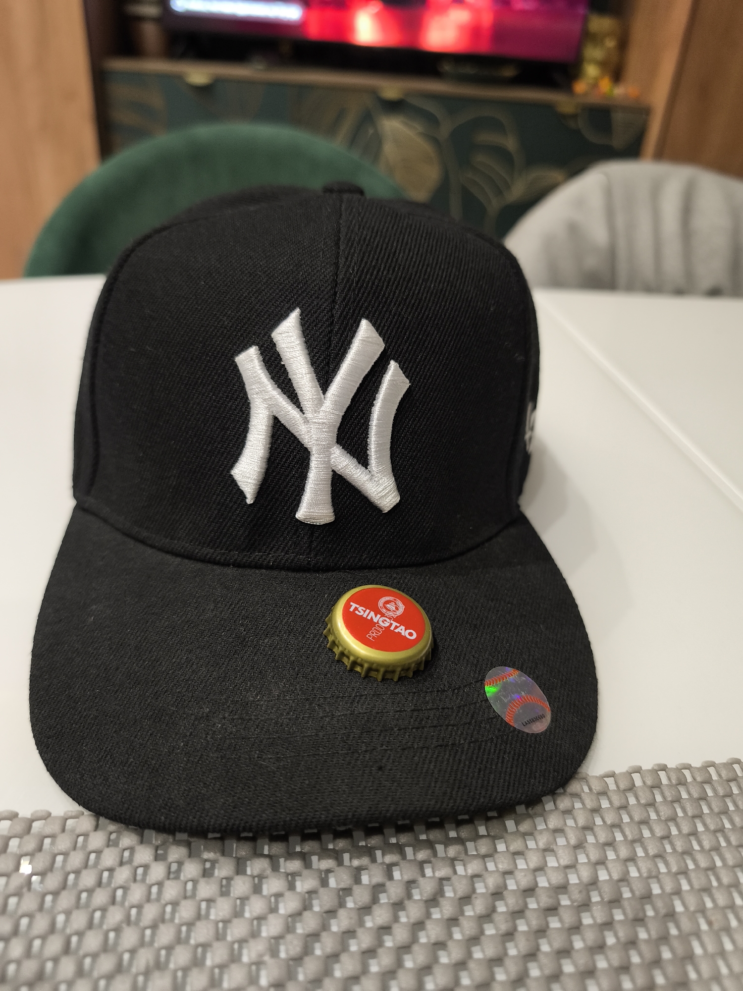 A black baseball cap with the New York Yankees logo is centered on a white surface. A bottle cap of Tsingtao beer rests on the brim of the cap, and a holographic baseball sticker is adhered near the edge of the brim. A gray placemat is partially visible at the bottom of the image. In the blurred background are elements of a home interior, including a wooden cabinet, a television displaying a red-toned image, and chairs upholstered in green and gray. The location in Moscow, Russia cannot be determined from the provided image.