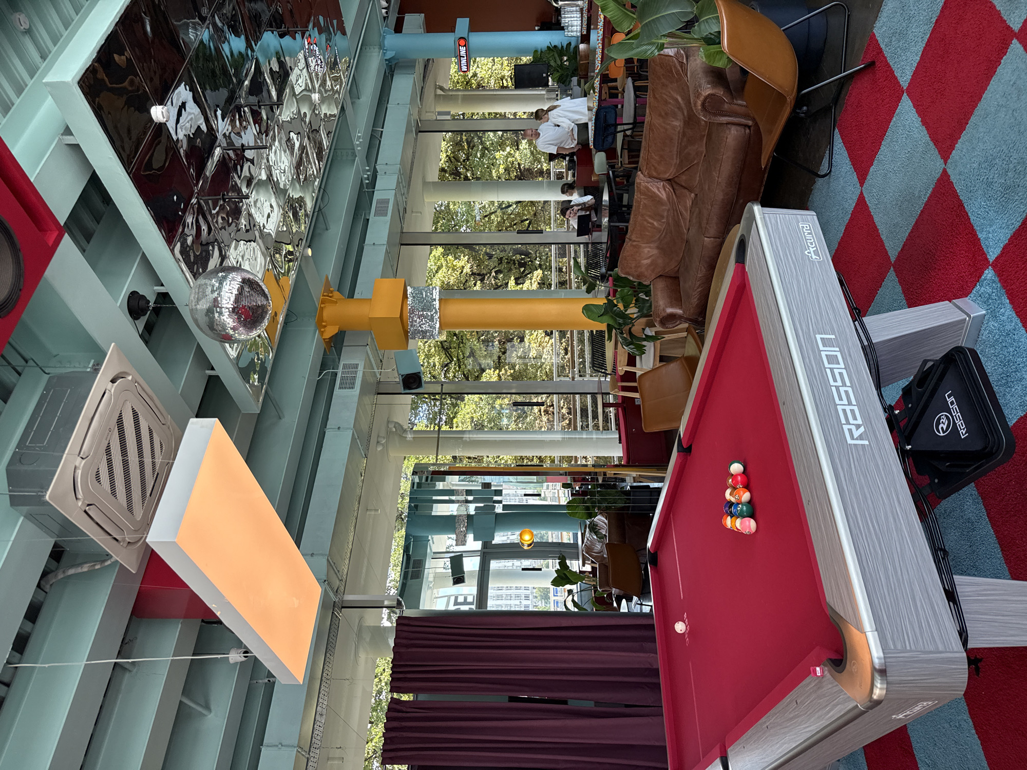 The image depicts a modern, well-lit indoor space, likely a bar or recreation area. In the foreground, a red pool table with a white cue ball and a rack of colored pool balls is prominently featured. The pool table has a "RASSON" logo. Behind the pool table, a brown leather sofa and a tan leather chair are visible. The room features large windows that offer a view of trees and buildings, suggesting an urban setting. The ceiling is industrial in style with exposed beams and pipes, and incorporates modern lighting fixtures and a disco ball. The floor is covered with a red and blue checkered carpet. Several people are present in the background, some standing and interacting, and one person appears to be working on a laptop at a table. There is also a bar area visible in the distance with drinks and bar stools. The overall atmosphere is contemporary and social. The presence of the "WINLINE" sign suggests the location is a betting or gaming establishment. Based on the "WINLINE" signage, this establishment is located in Moscow, Russia.