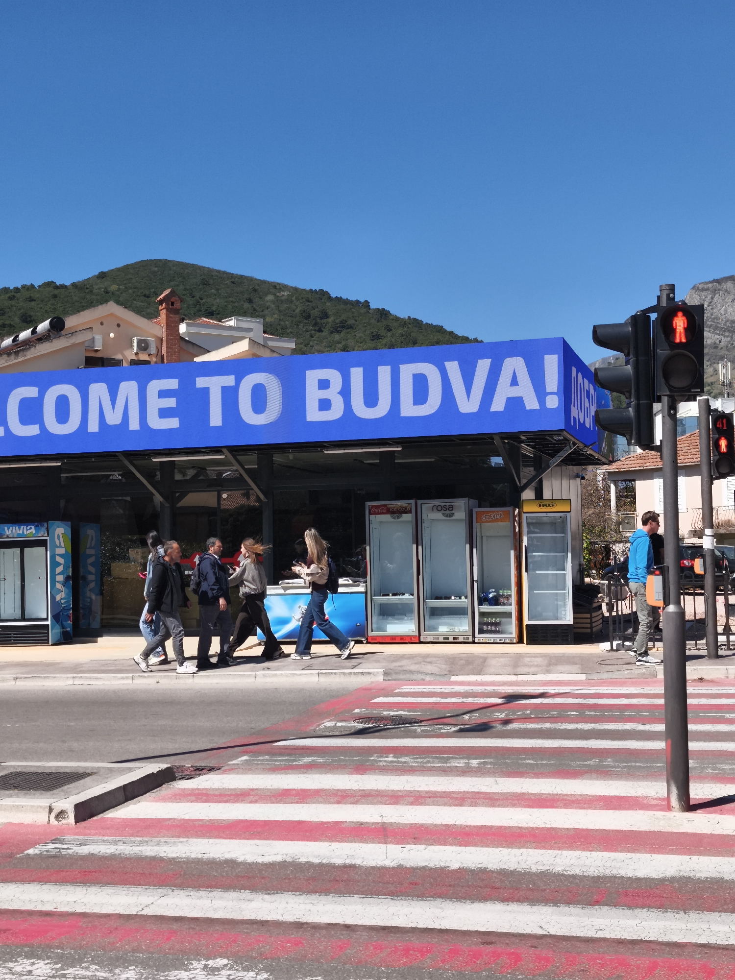 A street scene in Budva, Montenegro, shows a crosswalk with red and white stripes. A group of pedestrians, including two women, are crossing the street. Above them is a blue sign that reads, "WELCOME TO BUDVA!" The street is lined with vending machines. The traffic light is red for pedestrians. A man stands near the traffic light. In the background are buildings and a mountain. The sky is clear and blue.