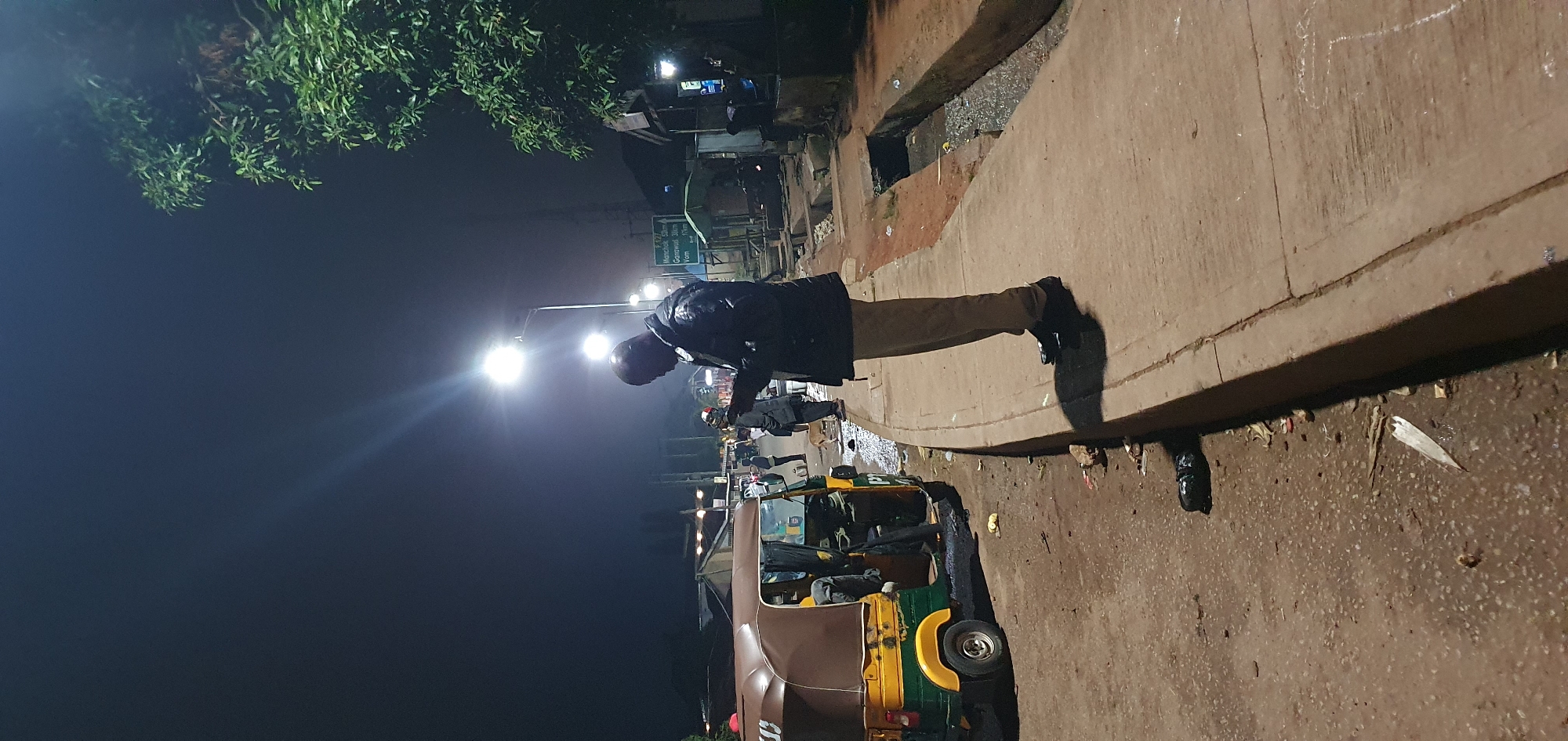 A full-shot, eye-level, vertical outdoor medium shot shows a man standing on a sidewalk looking at his phone at night in Bukuru, Nigeria. The man is in the center-right of the frame, wearing a dark jacket and light-colored pants. He is looking down at his phone, which he is holding in his hands. The sidewalk is made of concrete and is lit by streetlights. To the left of the sidewalk is a street with an auto-rickshaw parked on it. The auto-rickshaw is yellow and brown and has its doors open. There are other people in the background, as well as trees and buildings. The scene is illuminated by streetlights, casting long shadows on the ground. The ground is littered with trash, including plastic bags and debris.