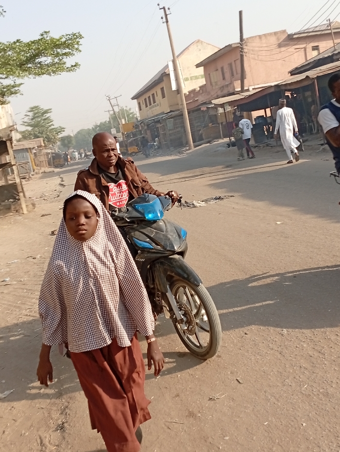 This image captures a street scene in Gama, Nigeria, featuring multiple individuals and structures under daylight conditions. In the immediate foreground, a young girl with dark skin walks forward, wearing a brown and white checkered long-sleeved top with a hood-like head covering and brown trousers; a light-colored bracelet is visible on her right wrist. Directly behind her, a bald, dark-skinned man in a brown jacket over a black t-shirt with a red 'Louis Vuitton' heart logo is positioned on a dark-colored LIFAN step-through motorcycle, his hands on the handlebars. The road surface is unpaved, dusty, and light brown. In the background, several one and two-story buildings line the street, some with corrugated metal roofs, suggesting commercial or residential use. Utility poles with overhead power lines are visible, crossing the frame. Other individuals are present further down the street, including a man in a white traditional robe walking from right to left, and another person wearing a white sports jersey with the number "9" on the back.