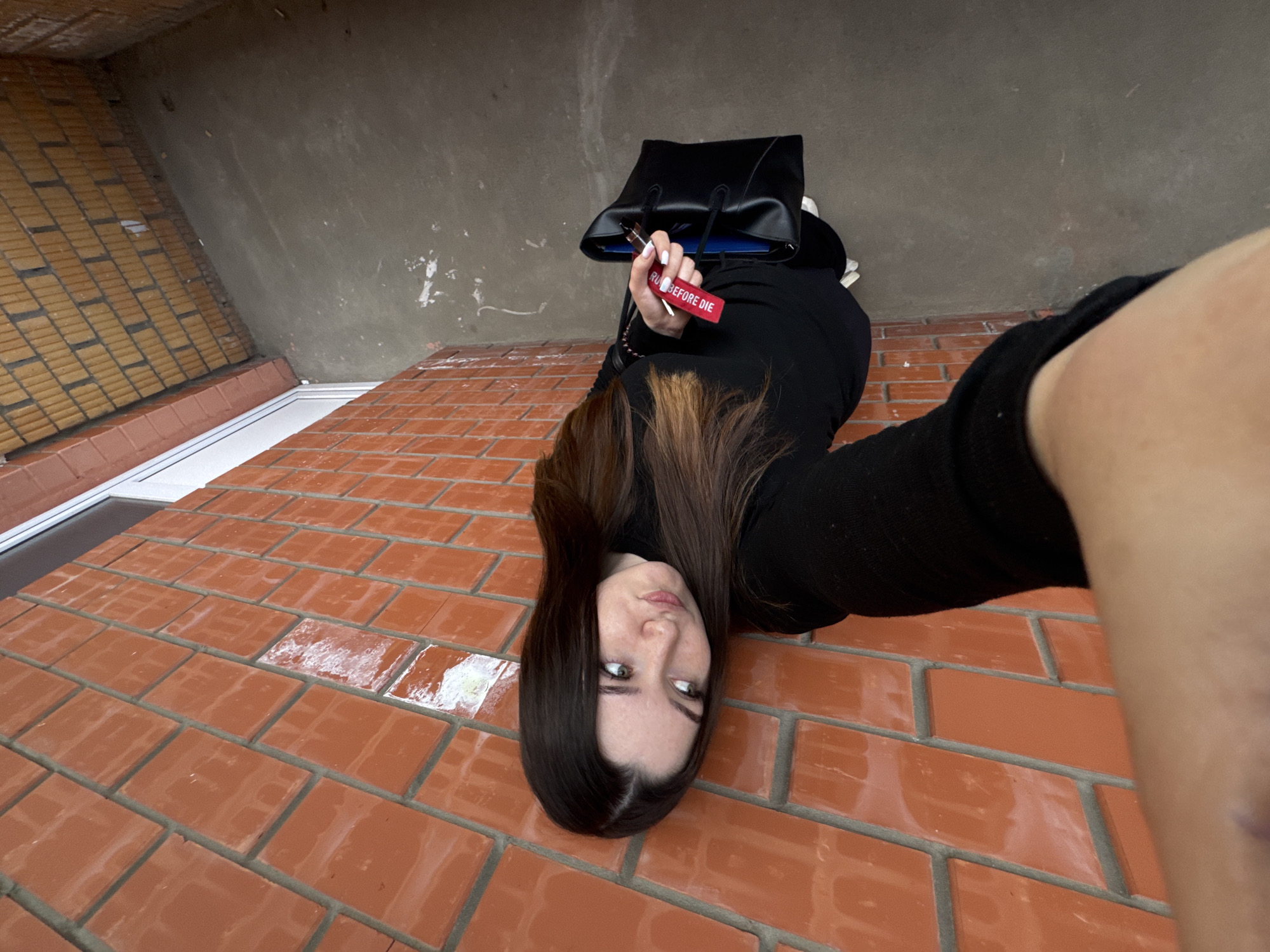 This media file shows a young woman lying on a brick surface, with her head facing downwards. She has long, dark brown hair and is wearing a black long-sleeved top. She is holding a black handbag with a red tag attached. The tag has white text that reads "RU? BEFORE DIE". The background consists of a grey textured wall and a section of yellow brickwork. The lighting suggests it is daytime, and the overall scene appears to be outdoors, possibly on a patio or walkway. The woman's expression is neutral. The location context is Perm, Russia.