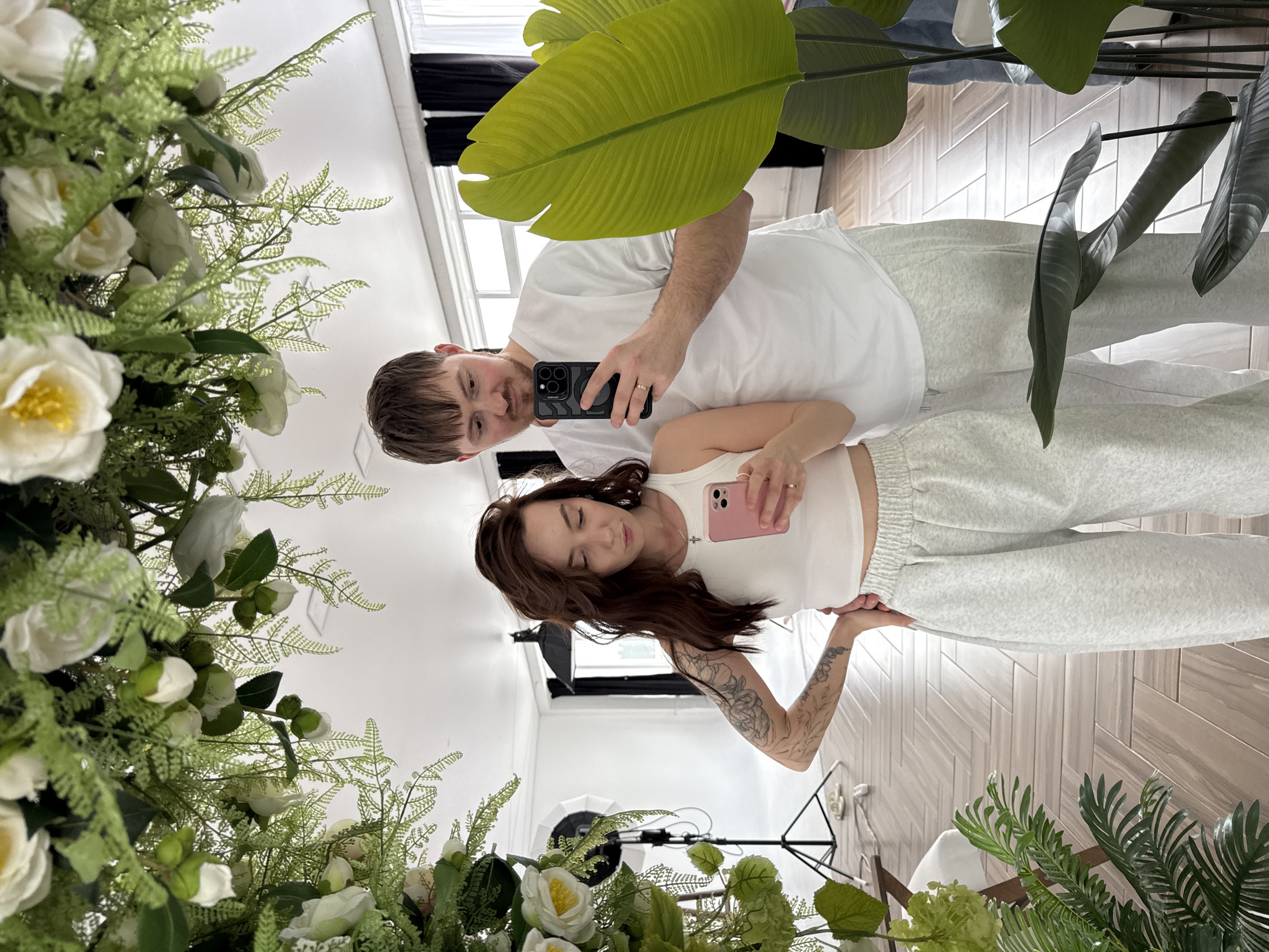 A man and a woman are taking a selfie in front of a mirror. The mirror is framed by floral arrangements and greenery. The man is holding a black iPhone, and the woman is holding a pink iPhone. Both are wearing casual clothing in light colors, and the woman has a tattoo on her left arm. In the reflection, studio lighting equipment is visible. The room has herringbone wood flooring. Based on the information provided, this image could potentially be in Chaikovsky, Russia.