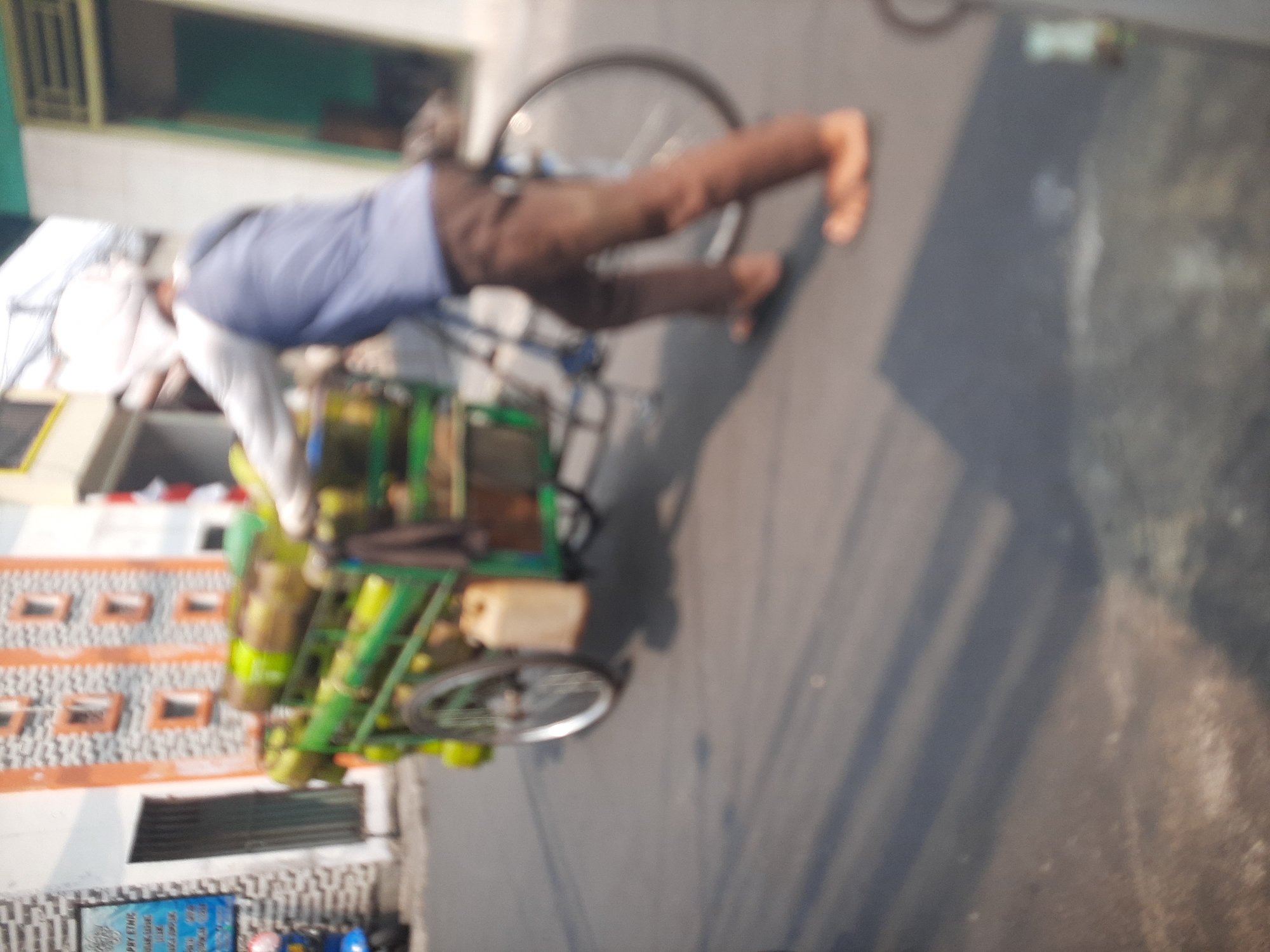 In a city scene in Central Jakarta, Indonesia, a person is pushing a bicycle cart loaded with green cylinders, likely gas canisters. The cart has one visible wheel, a yellow jerrycan, and is positioned on a paved street. The person is wearing a blue long-sleeved shirt, brown pants, and a white head covering. They are actively engaged in moving the cart forward. The background shows buildings with distinctive orange-trimmed windows and a wall with a textured facade. A blue and white sign is also visible on the left side of the frame. The overall image has a blurred quality, suggesting movement or a low-resolution photograph. The lighting indicates it is daytime with shadows cast on the street.