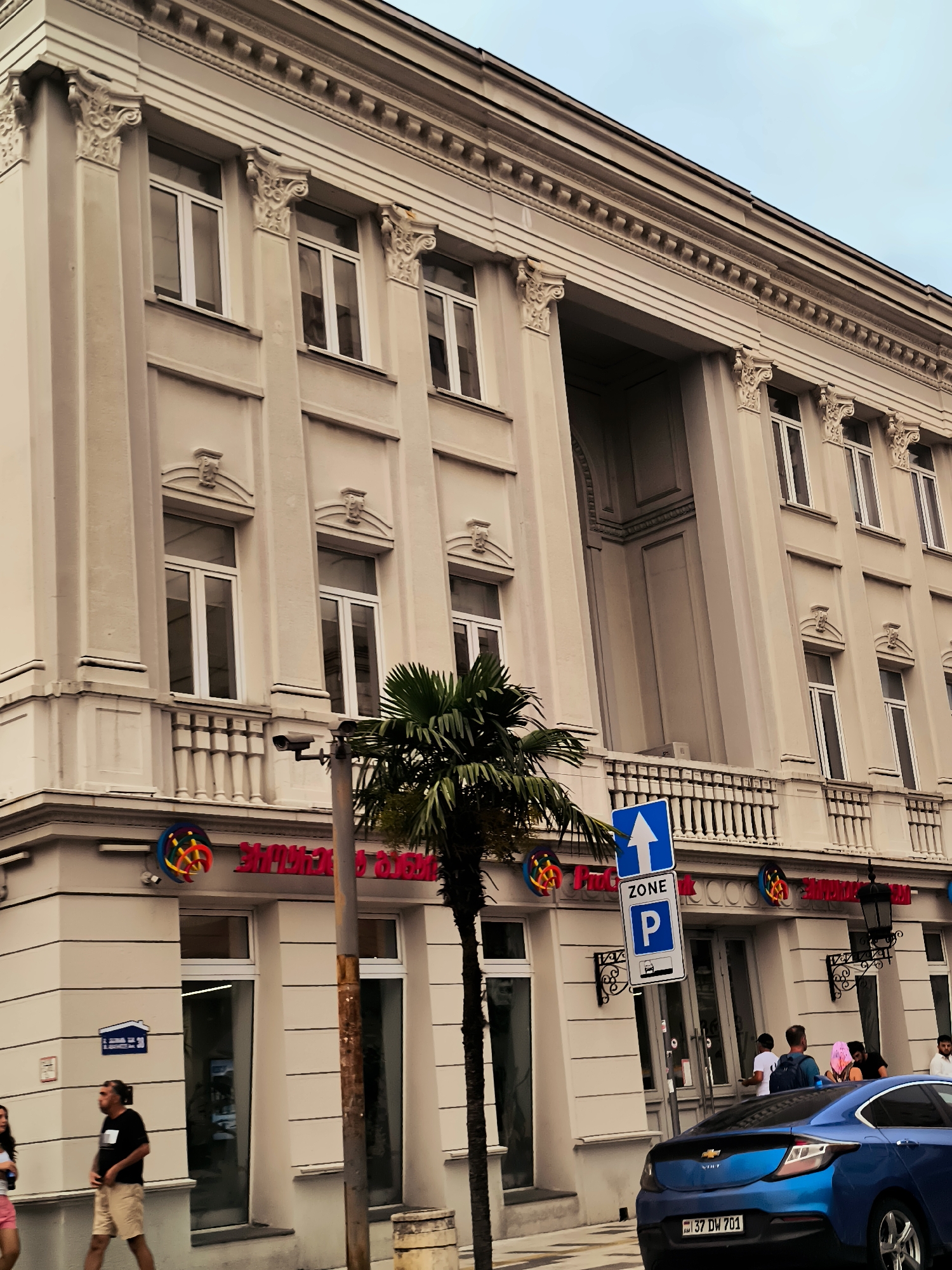 A building with classical architectural elements, featuring a beige facade, columns with ornate capitals, and numerous windows. On the ground floor, there are storefronts with large glass windows and red lettering on signage. A palm tree stands in front of the building. A blue Chevrolet Volt is parked on the street. Several people are visible on the sidewalk and near the storefronts. A parking zone sign with an upward arrow is prominently displayed. The building appears to be located on a street in Batumi, Georgia, indicated by the Georgian license plate on the car.