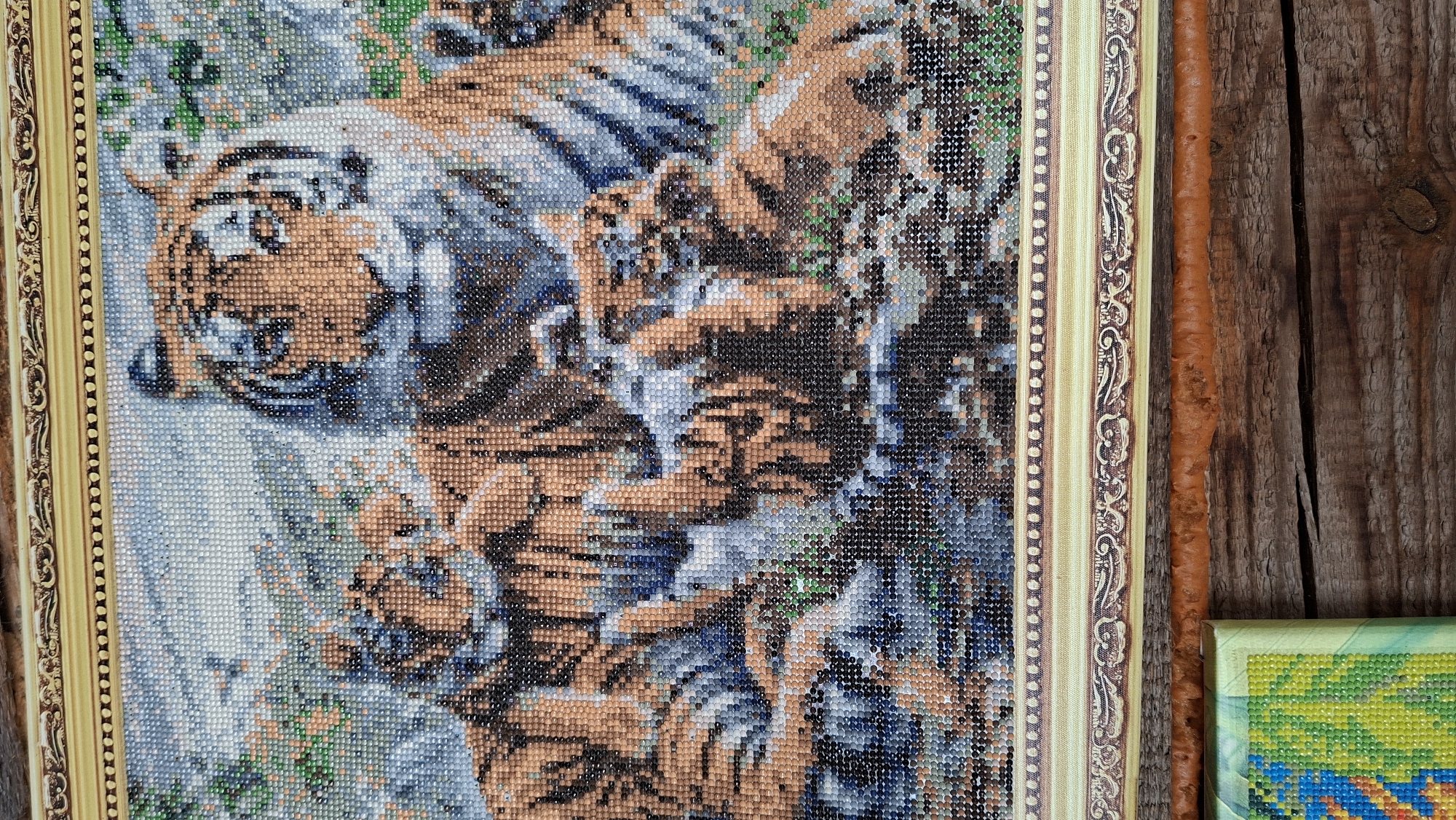 The image shows a framed diamond painting depicting tigers. The painting has a gold-colored frame with decorative patterns. The background is made of wooden planks. Another diamond painting, partially visible, is located on the right-hand side of the frame. The photograph might be taken in a domestic setting. Location in the city of Большой Букор, Russia, cannot be confirmed.