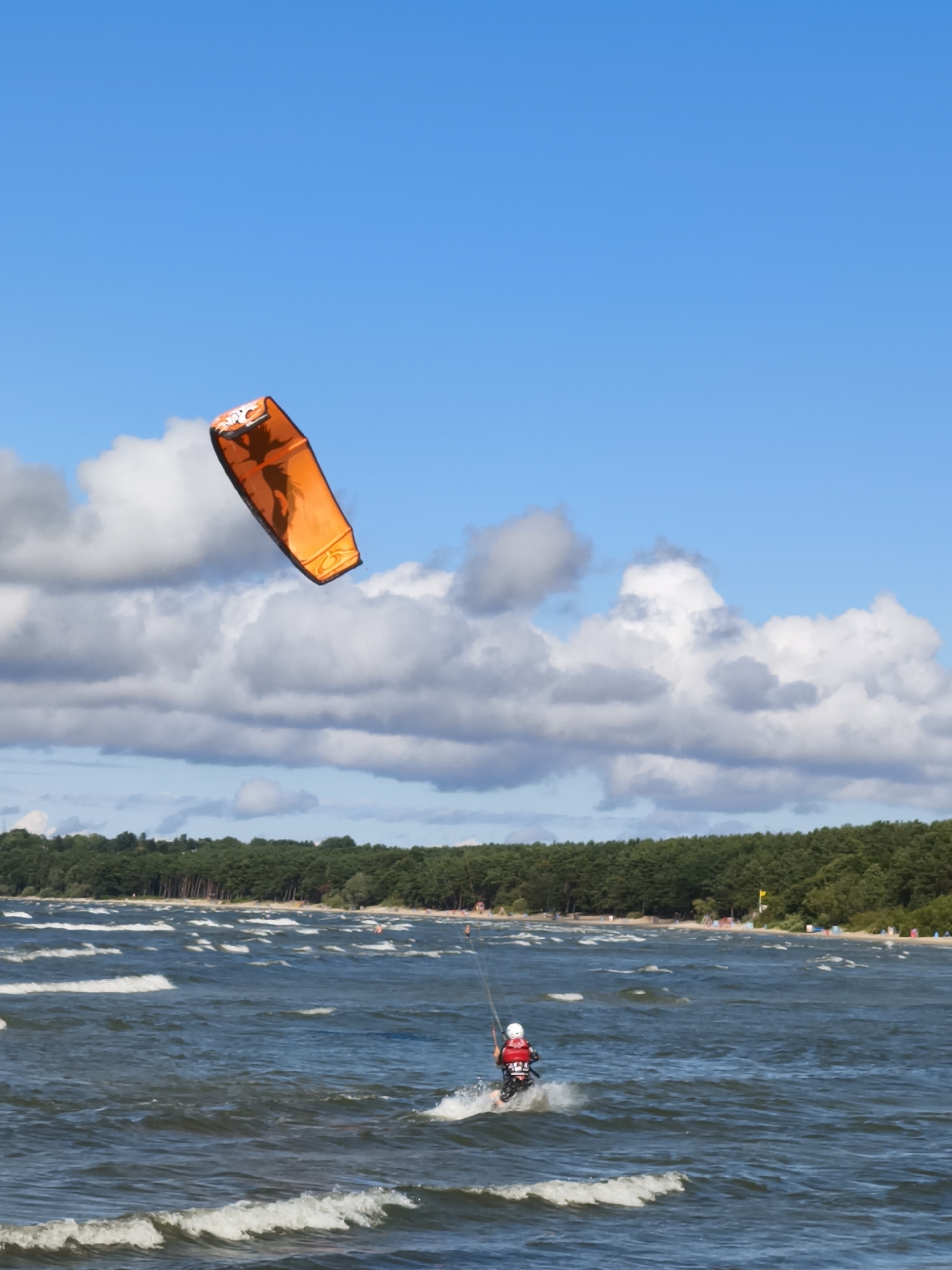 An orange kite is visible in the sky above a person on a surfboard, kiteboarding in the sea. The person is wearing a red helmet and jacket. The sea is choppy, with waves and whitecaps. In the background, there is a beach lined with trees. The sky is blue with some clouds.