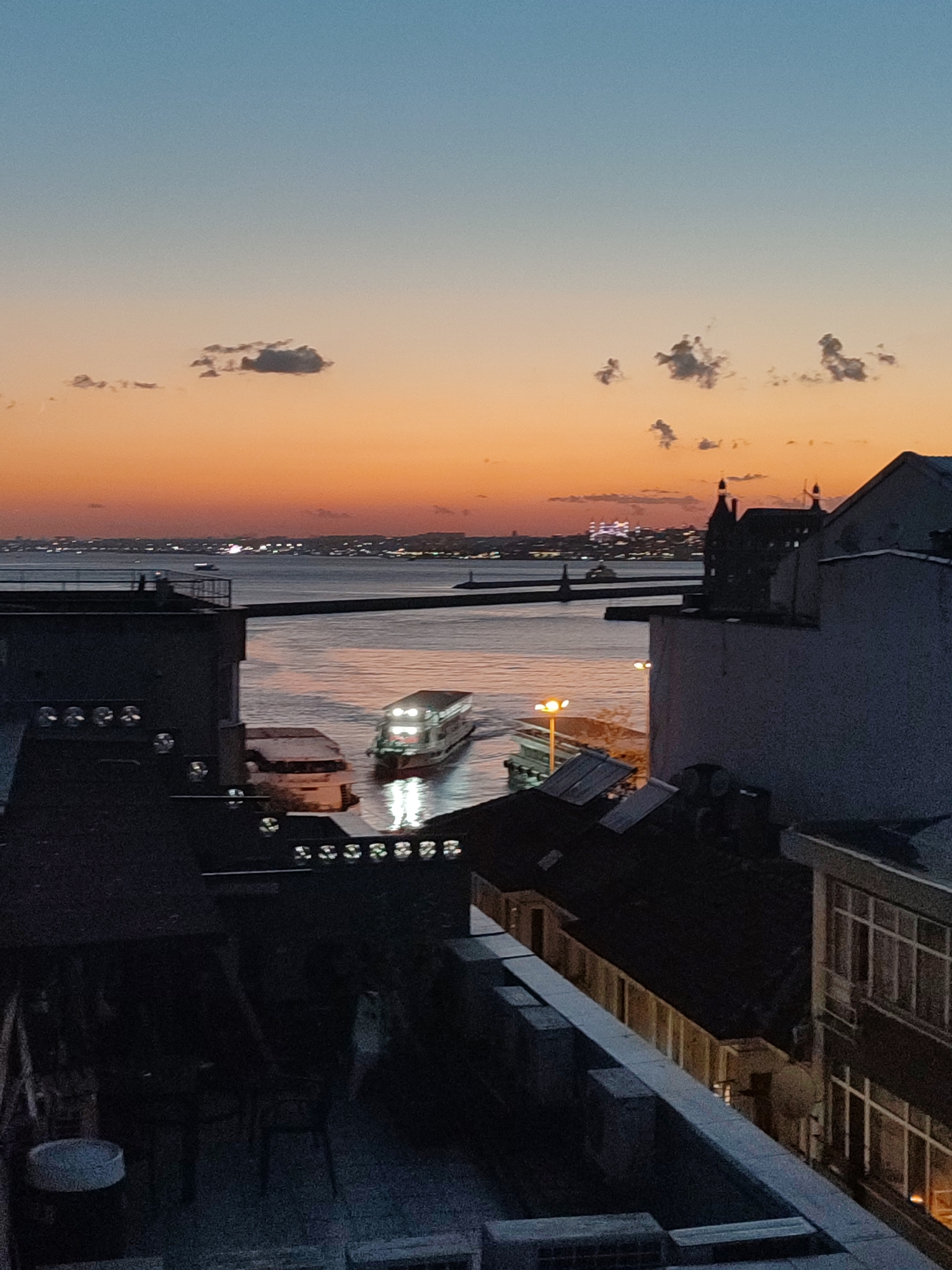 The image depicts a dusky waterfront scene in Kadıköy, Turkey, with a striking sunset painting the sky in hues of orange and blue. In the foreground, the dark silhouettes of buildings and rooftops dominate, suggesting an urban setting. A ferry, illuminated with lights, cruises on the water, its reflection shimmering on the surface. In the distance, the city skyline is visible, dotted with lights, and a few clouds drift across the horizon. The overall atmosphere is serene and tranquil, capturing a moment of transition from day to night in a vibrant coastal city.