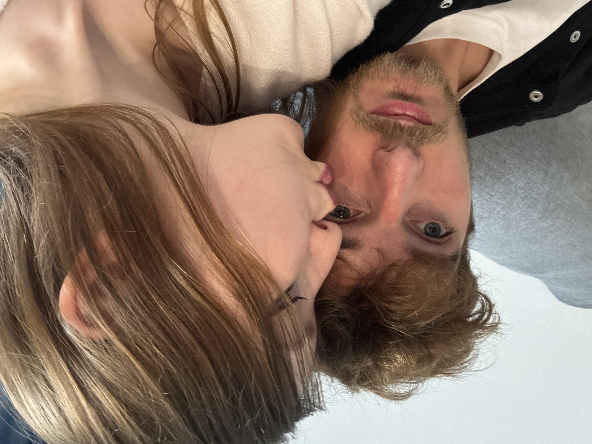 A close-up, waist-up selfie shows a young man and a young woman. The man, with light brown, messy hair and a light beard, is on the left side of the frame, looking at the camera. He has blue eyes and is wearing a white t-shirt under a black jacket with metal snaps. The woman is on the right side of the frame, leaning in to kiss the man on the cheek. She has long, light brown hair and is wearing a cream-colored textured sweater. The background is a neutral wall.