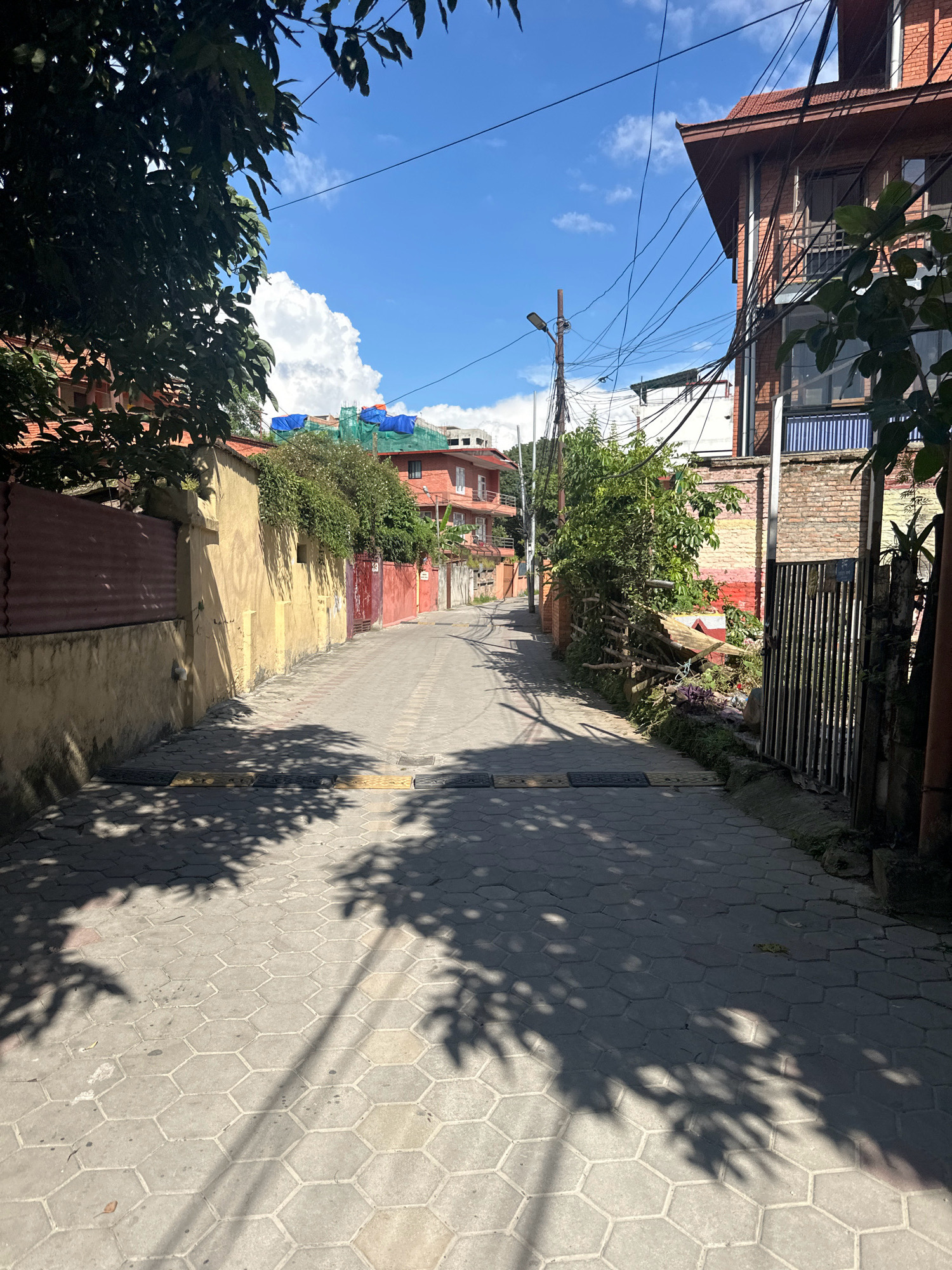 The image shows a narrow street in Lalitpur, Nepal. The street is paved with hexagonal concrete tiles and has speed bumps. On the left side of the street, there is a yellow plastered wall with a corrugated metal fence. On the right side, there are brick buildings with a metal gate and some overgrown vegetation. Overhead, a tangle of electrical wires runs between poles. The sky is blue with some white clouds. The scene appears to be in a residential or commercial area.