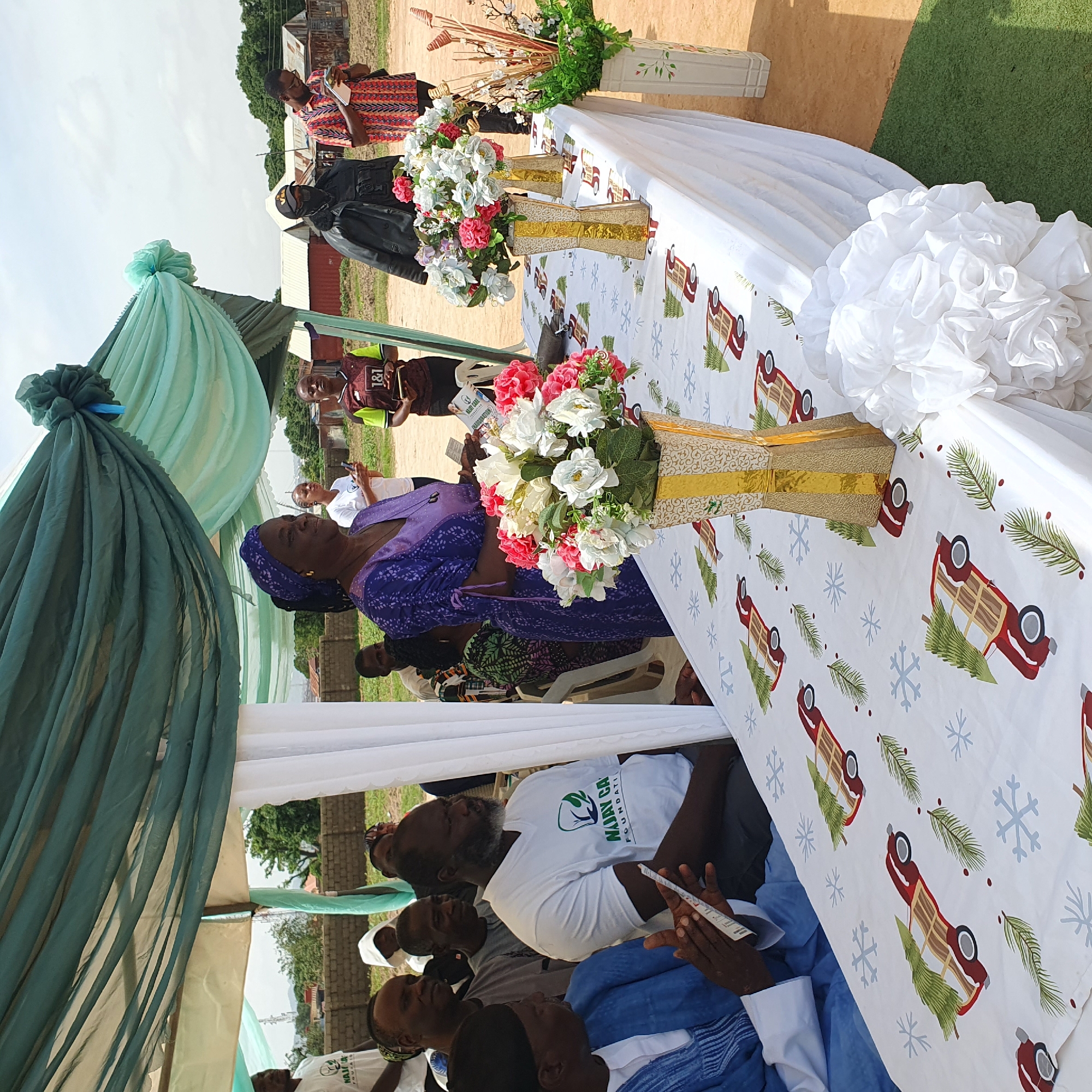 The image shows an outdoor gathering in Unguwar Saya, Nigeria. A group of people are seated under a tent with light blue and green drapes. A long table is covered with a white tablecloth adorned with a festive pattern of red trucks, Christmas trees, and snowflakes. Floral arrangements in vases are placed on the table.
Several individuals are visible in the foreground and background. A woman in a purple outfit and headscarf is looking towards the camera. A man in a white t-shirt with green text is also prominent. Other people are seated, some looking towards the table or engaged in conversation.
In the background, there are buildings, some appearing to be made of corrugated metal. Trees and a clear sky suggest it is daytime, possibly a sunny day. The overall atmosphere appears to be that of a community event or celebration.
Visible text on the t-shirt reads "MAJAY CA FOUNDATION".