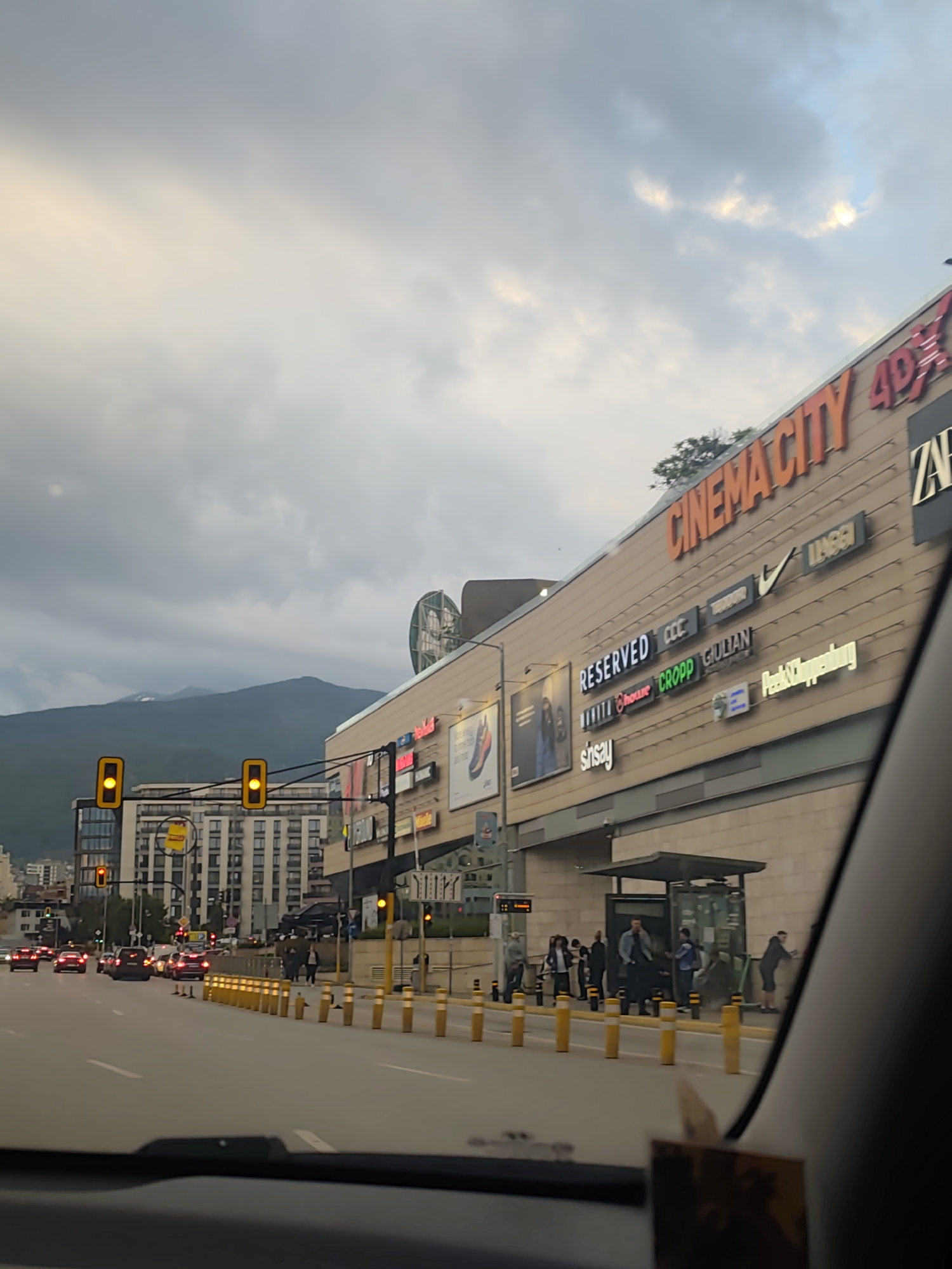 This image depicts an urban street scene in Sofia, Bulgaria, captured from inside a vehicle, with a portion of the dashboard visible at the bottom. The main subject is a large, multi-story commercial building on the right, which appears to be a shopping center and cinema complex, featuring a light brown facade.
The building's exterior is adorned with numerous illuminated brand signs. The most prominent, in orange, reads "CINEMACITY 4DX." Below it and to the right, part of a "ZARA" sign is visible. Other clearly readable brand names include "RESERVED," "CCC," "CROPP," "GIULIAN," "sinsay," the Nike swoosh logo, and "LIU JO," along with a partially visible "Peek & Cloppenburg" sign. People are seen congregating near the entrance or bus stop area of the building.
In the foreground, a multi-lane road is filled with several cars, many of which have their red brake lights illuminated, suggesting either stopped traffic at an intersection or slow movement. Two bright yellow traffic lights are conspicuous on the left side of the road. A line of yellow bollards separates the roadway from the sidewalk.
Behind the immediate street level, other urban buildings with modern architecture are visible. In the distant background, majestic mountains rise, partially obscured by heavy, grey clouds. These are characteristic of the Vitosha mountain range.
The sky is predominantly overcast, with some lighter, brighter patches indicating sunlight breaking through the clouds, suggesting either late afternoon or early evening. The artificial illumination from the building signs and traffic lights reinforces this time of day. The scene conveys a sense of a busy city environment.