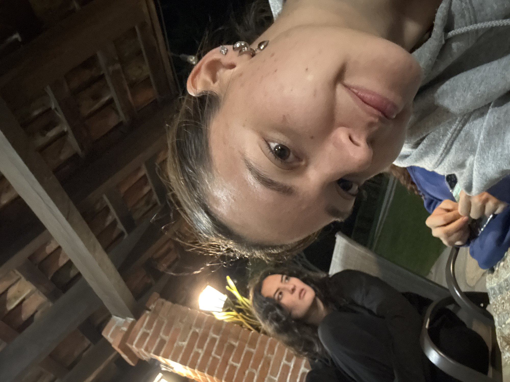 The photograph captures a dimly lit outdoor scene, possibly in the evening. Two women are visible. One, closer to the camera, wears a grey hooded sweatshirt and has multiple piercings in her left ear. The other woman, positioned further back and slightly below, is wearing a dark shirt. In the background, a red brick column supports a wooden structure, likely part of a building or covered patio. The setting appears to be a casual outdoor gathering. The location in the city of Puebla, Mexico cannot be determined from the image.