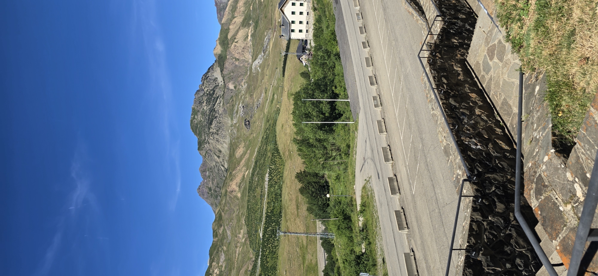 This is a landscape photograph of a mountainous region on a clear, sunny day. The image is oriented vertically, with the sky occupying the left side and the mountainous terrain and a paved area on the right. In the background, a large, multi-story building with a light-colored facade and dark windows is visible on a grassy slope. Below the building, a winding paved road or parking lot is lined with concrete barriers. Beyond the road, a verdant mountainside slopes upwards, dotted with patches of trees and vegetation. The distant mountains are rugged and rocky, with varying shades of brown and green. A metal tower with crisscrossing supports is visible in the mid-ground. The foreground features a stone retaining wall with a metal railing, looking down onto a shaded area with more stone and grass. The overall scene suggests a location in a mountainous resort or alpine area, likely during the daytime given the bright sunlight and clear blue sky with a few wispy clouds. There is no visible text in the image.