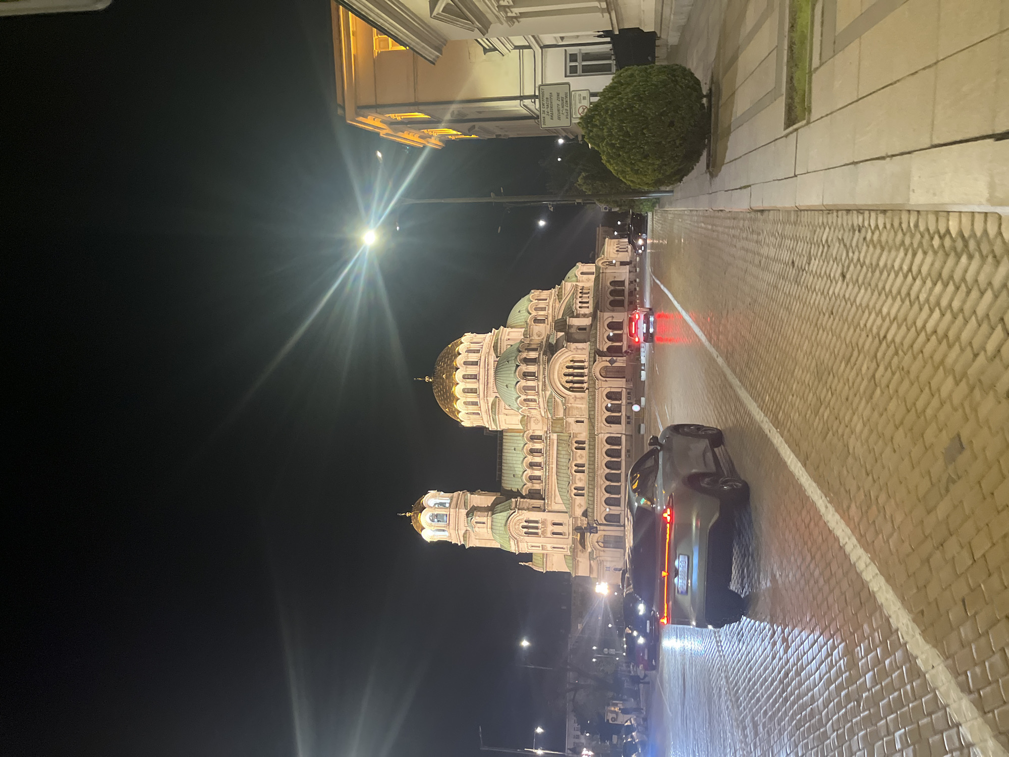 The image displays a nighttime street scene in Sofia, Bulgaria, featuring the illuminated Alexander Nevsky Cathedral. The cathedral, a grand and ornate structure, dominates the right half of the frame with its numerous domes, some adorned with golden gilding and others with green patinas. Its intricate architectural details, including arched windows and decorative elements, are highlighted by warm lighting.
A cobblestone street runs through the foreground and recedes into the distance, bordered by a paved sidewalk on the left. A dark gray car is visible in the mid-ground, its red taillights glowing, and another red vehicle is further down the street, along with other distant car lights. Bright streetlights punctuate the dark sky, causing visible lens flares and casting illumination on the street and buildings.
On the left side, other classical buildings are partially visible, also lit. A spherical, green bush stands on the sidewalk. A white sign attached to a pole displays text in Cyrillic, followed by "SECURITY ZONE" and "VIDEO CONTROL" in English. The scene depicts a serene urban evening around a magnificent historical landmark.