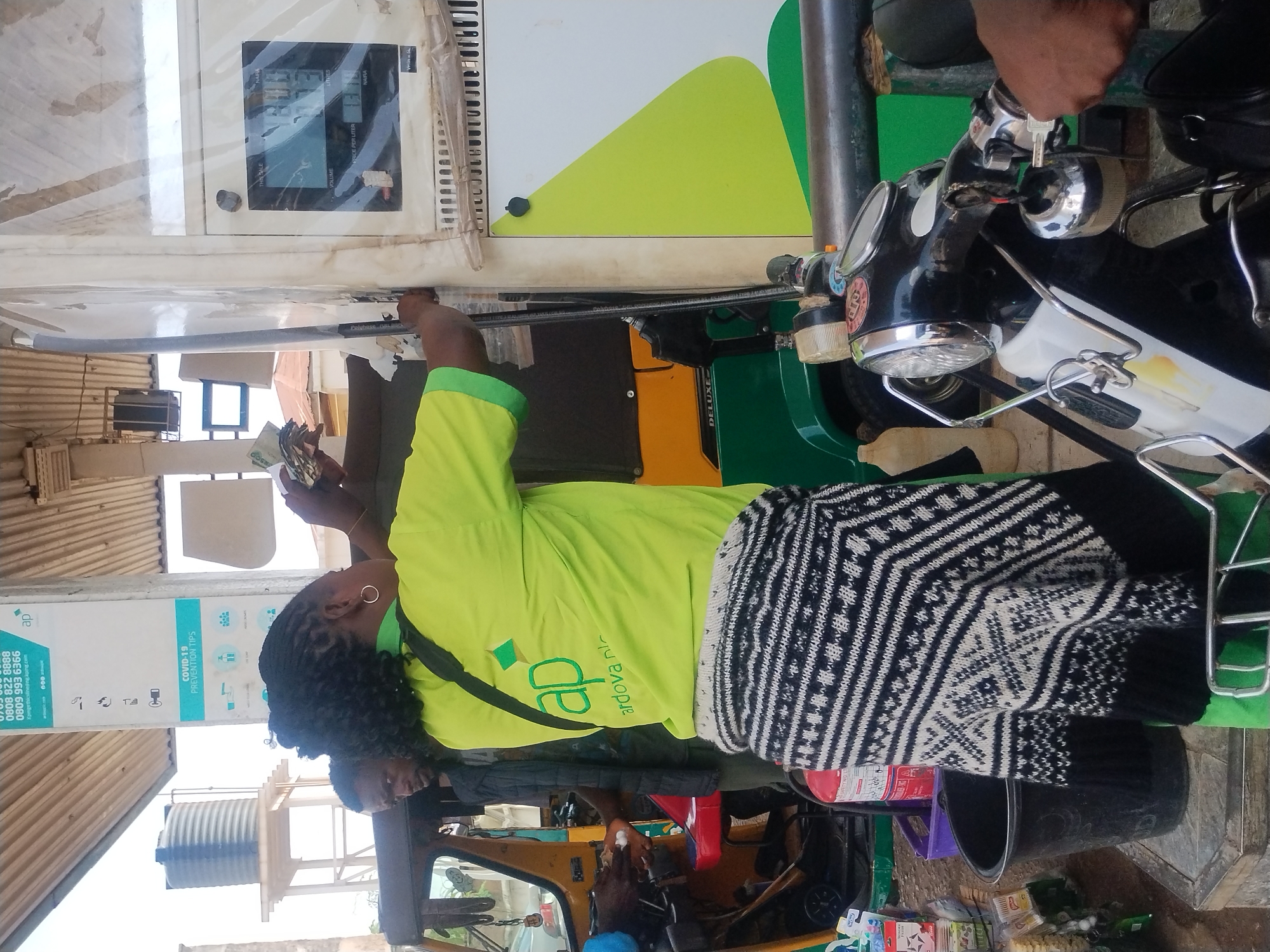 In Jos, Nigeria, a woman in a bright green shirt with a black and white patterned skirt is handing cash to an attendant at a gas station. The attendant, a man with dark skin and braided hair, is wearing a black jacket and is holding cash in his left hand while operating the fuel pump with his right. Behind them, a yellow and green auto-rickshaw is parked, its driver visible in the background. A black motorcycle with chrome accents is in the foreground. The gas station has a white and green structure with a visible fuel pump display showing numbers. Various items, including bottles and containers, are scattered around the station. The overall scene depicts a transaction at a local gas station.
