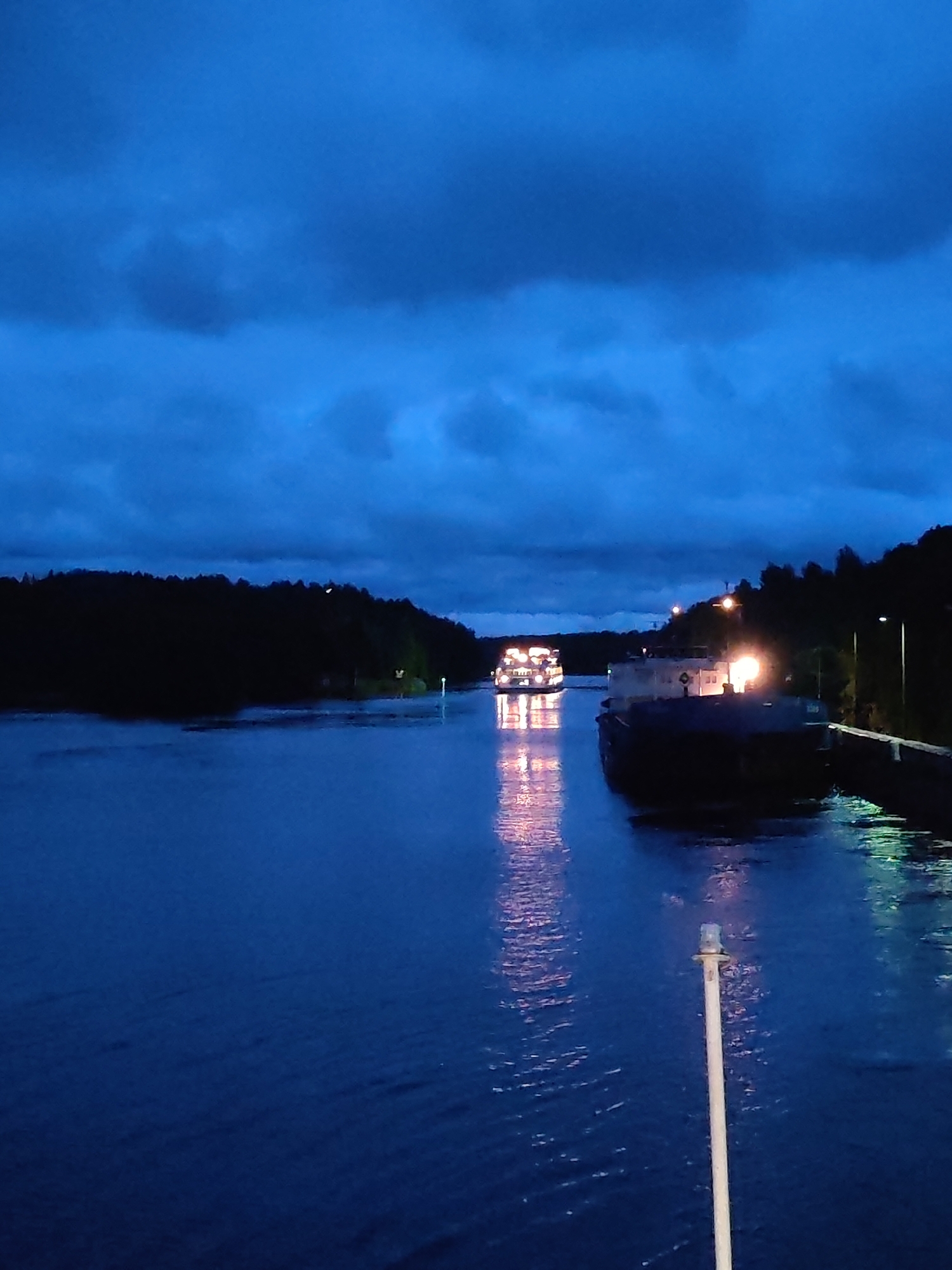At dusk in Devyatiny, Russia, two cargo ships navigate a dark canal bordered by a dense, tree-covered shoreline. The sky is a deep blue, filled with ominous clouds. The water reflects the lights of the ships, creating shimmering trails. The lead ship is brightly lit, its windows glowing warmly, while the second ship, closer to the foreground, is partially obscured by darkness but also emits a warm light from its deck. The canal walls are visible on the right, with streetlights casting pools of illumination. A tall, slender pole with a light at its top stands in the foreground, its reflection stretching across the water. The scene conveys a sense of peaceful solitude, with the ships moving purposefully through the quiet evening.