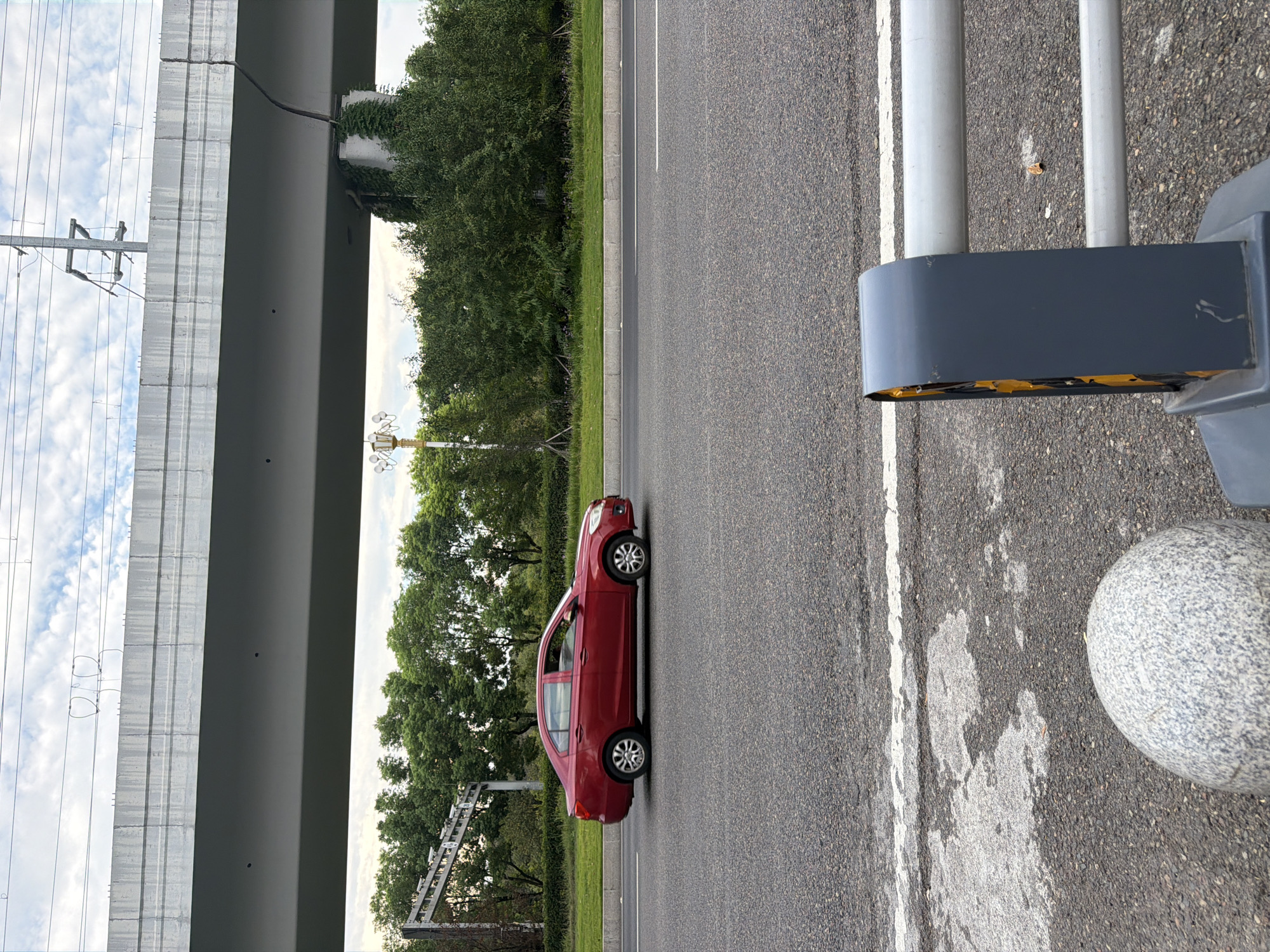 A red sedan is driving on a multi-lane asphalt road, passing beneath a concrete overpass. The overpass has electrical wires and supports visible. On the side of the road, there are green trees and a grassy median. In the foreground, a grey granite bollard and a section of a grey metal barrier with reflective orange elements are visible. The sky above is partly cloudy. The scene is consistent with an urban environment in Jiangjin, China, likely near a highway or elevated roadway.